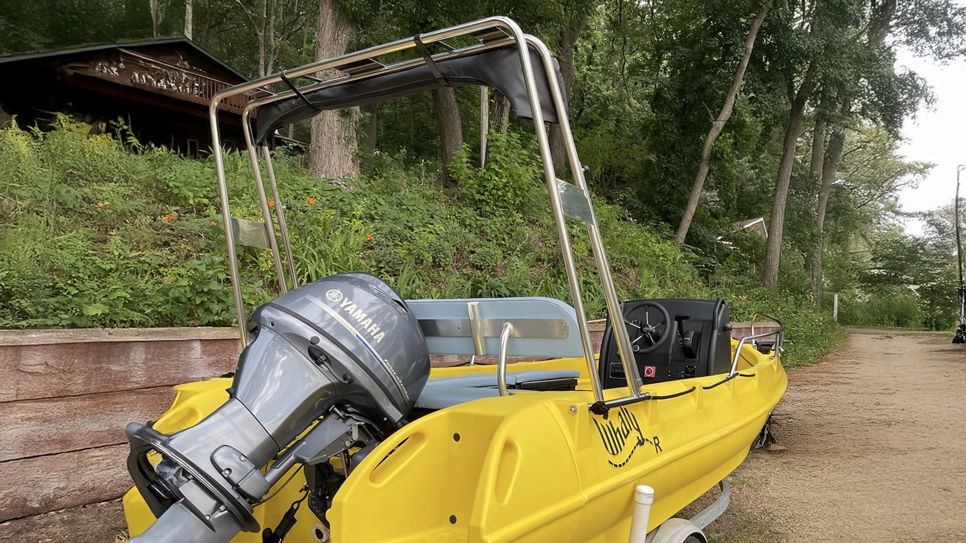 Yellow motorboat with Yamaha outboard motor on a trailer, parked on a dirt path near a wooden landscape wall and green trees.