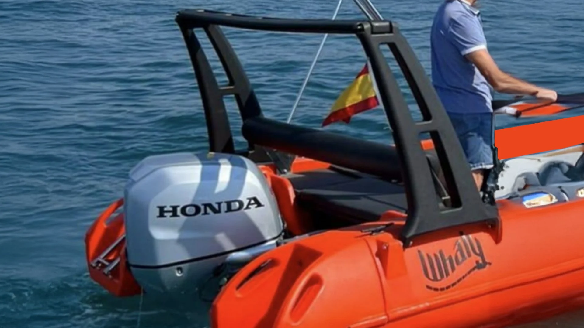 Orange inflatable boat with a Honda outboard motor in the water, a person in a blue shirt standing on the boat, and a small flag attached to the boat.
