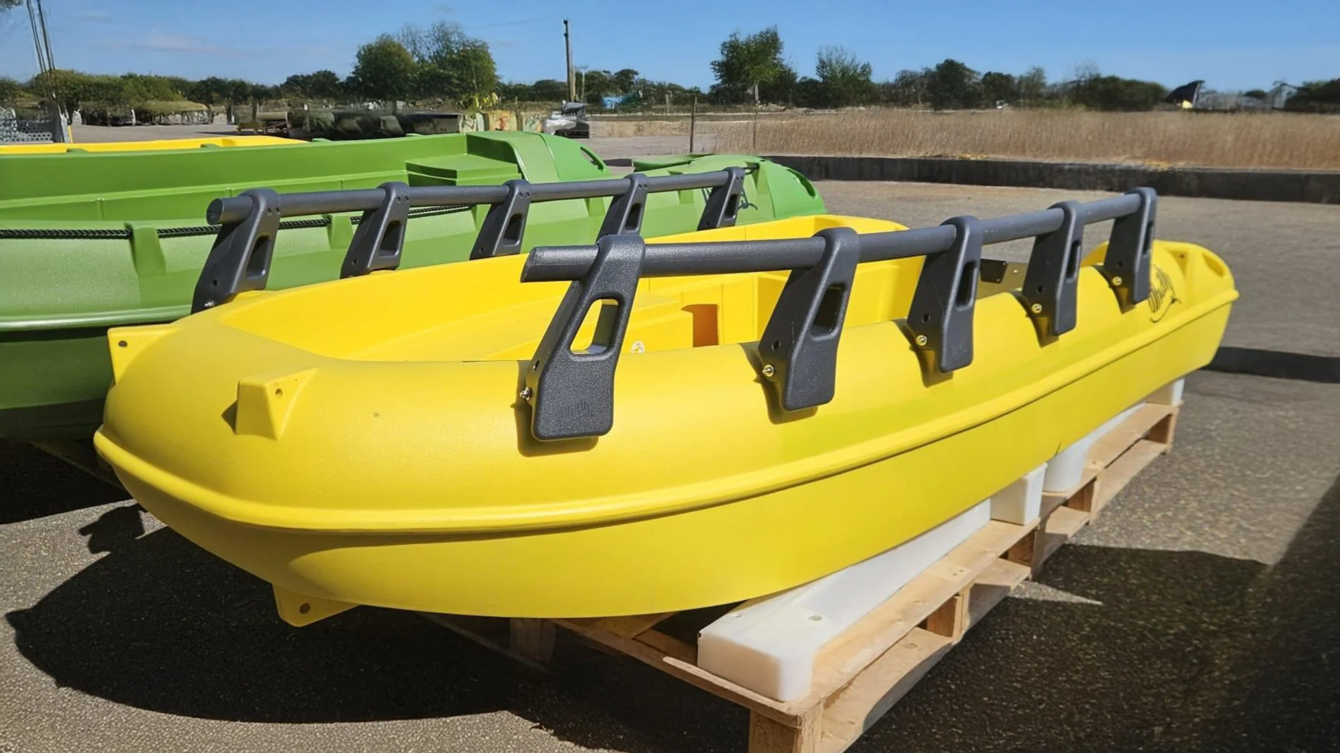 A yellow rescue boat with black handles on a wooden pallet outdoors, with a green boat behind it and a clear blue sky in the background.