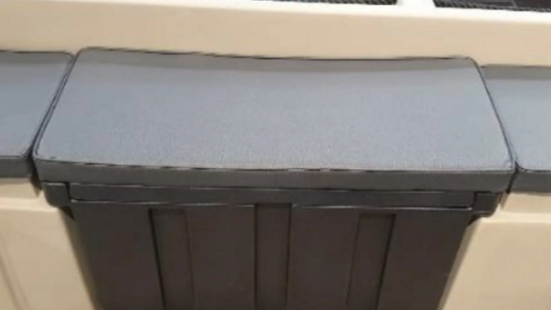 Close-up of a black and gray trash or storage bin with a lid, mounted against a beige wall.