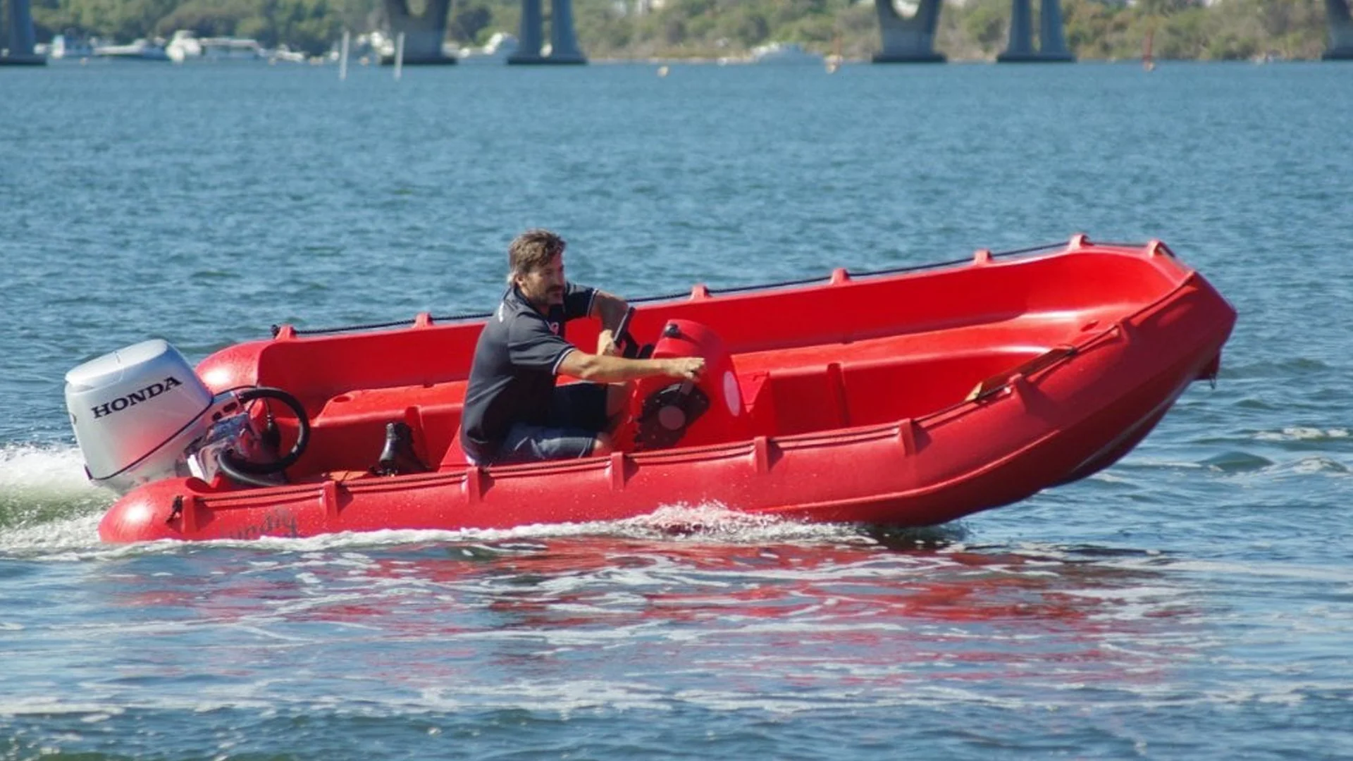 Man in black shirt driving a small red boat with a Honda outboard motor on a body of water.