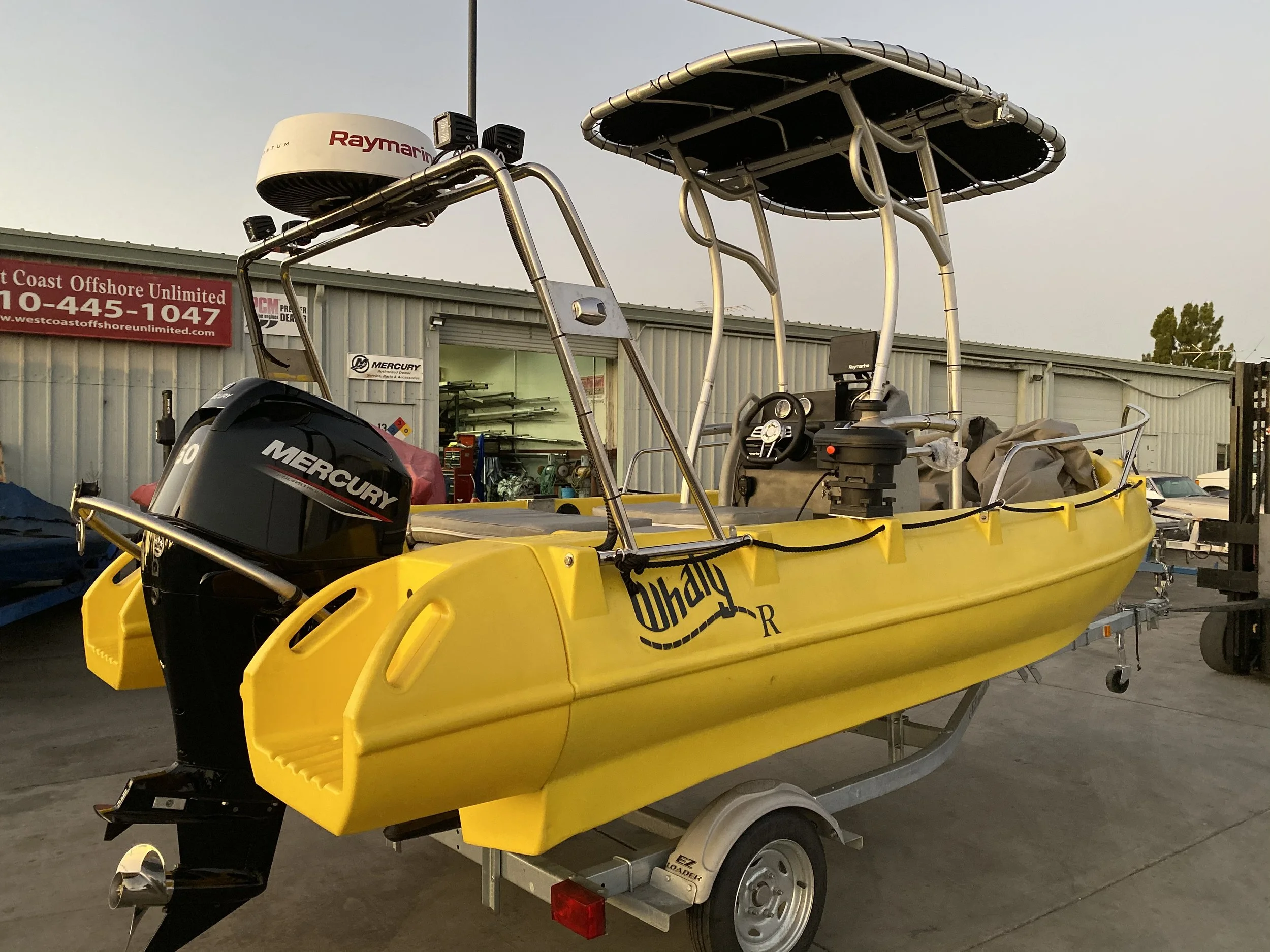 Yellow 500R Whaly boat on a trailer, equipped with a Mercury outboard motor and Raymarine radar, parked in an outdoor area near a building with signs and tools.