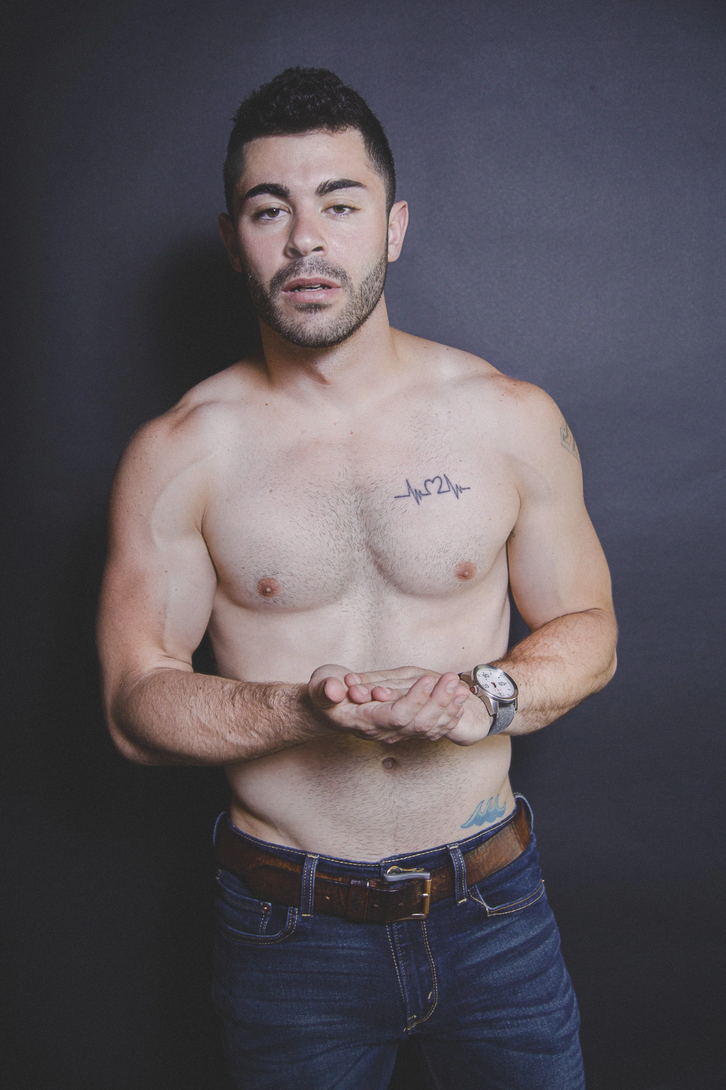 A shirtless man with tattoos, wearing jeans and a watch, posing against a dark background.