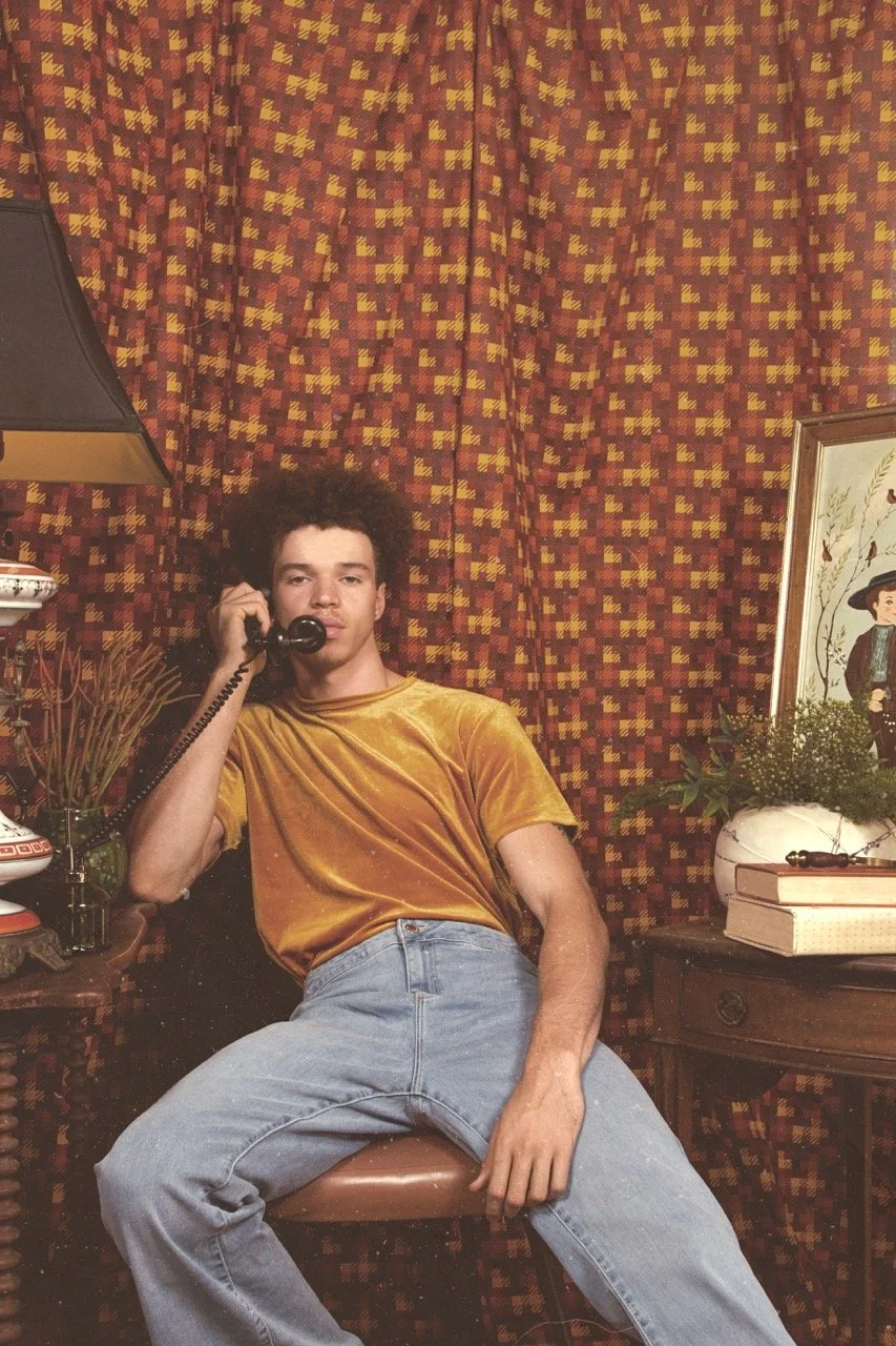 A young man with curly hair wearing a yellow velvet T-shirt and light blue jeans sitting on a chair, talking on a corded telephone in a room with a red and yellow patterned curtain background. There are decorative items on the side tables including a
