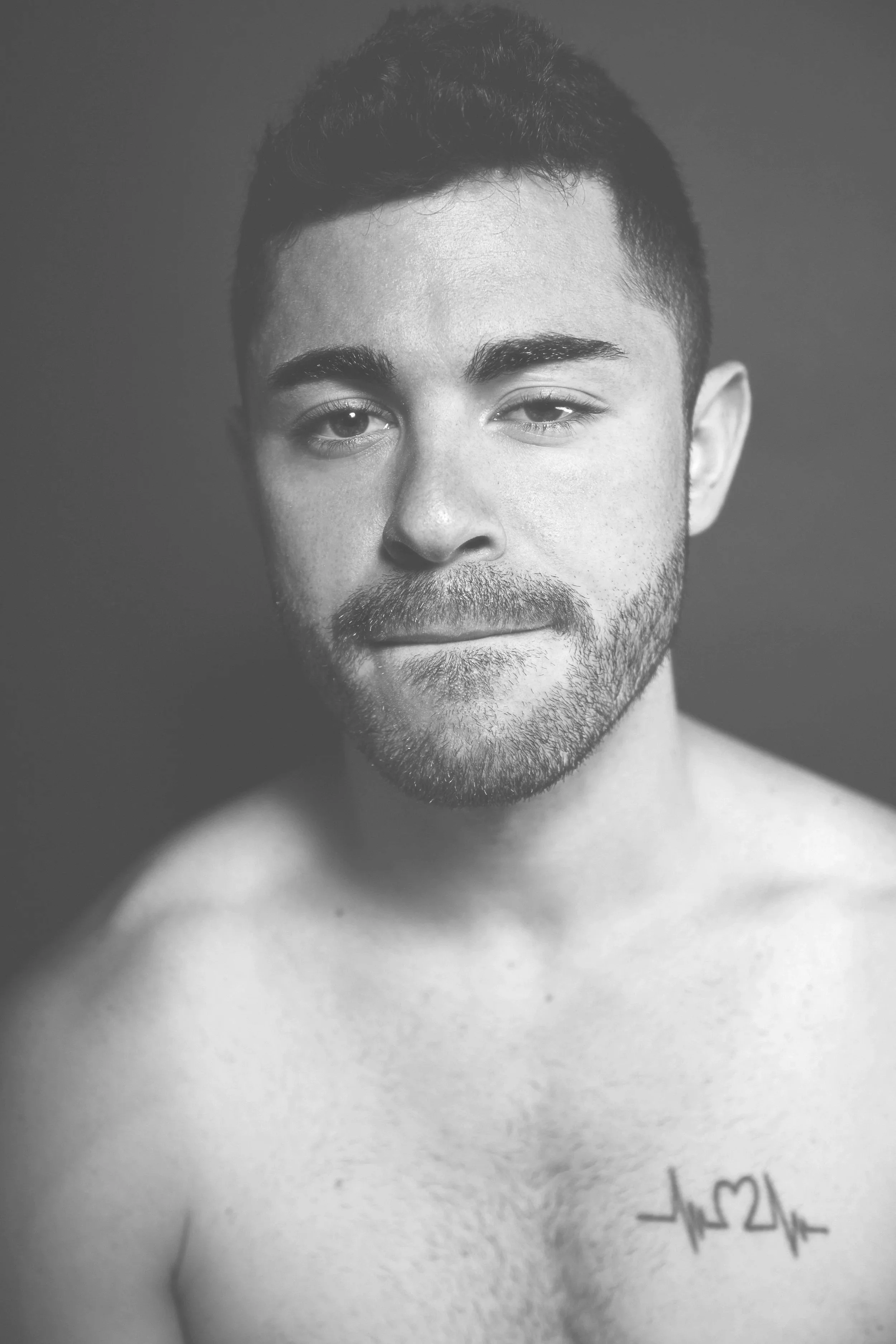 Black and white portrait of a young man with short hair and a beard, looking at the camera with a slight smile, shirtless, with a tattoo of a heartbeat line and heart on his chest.