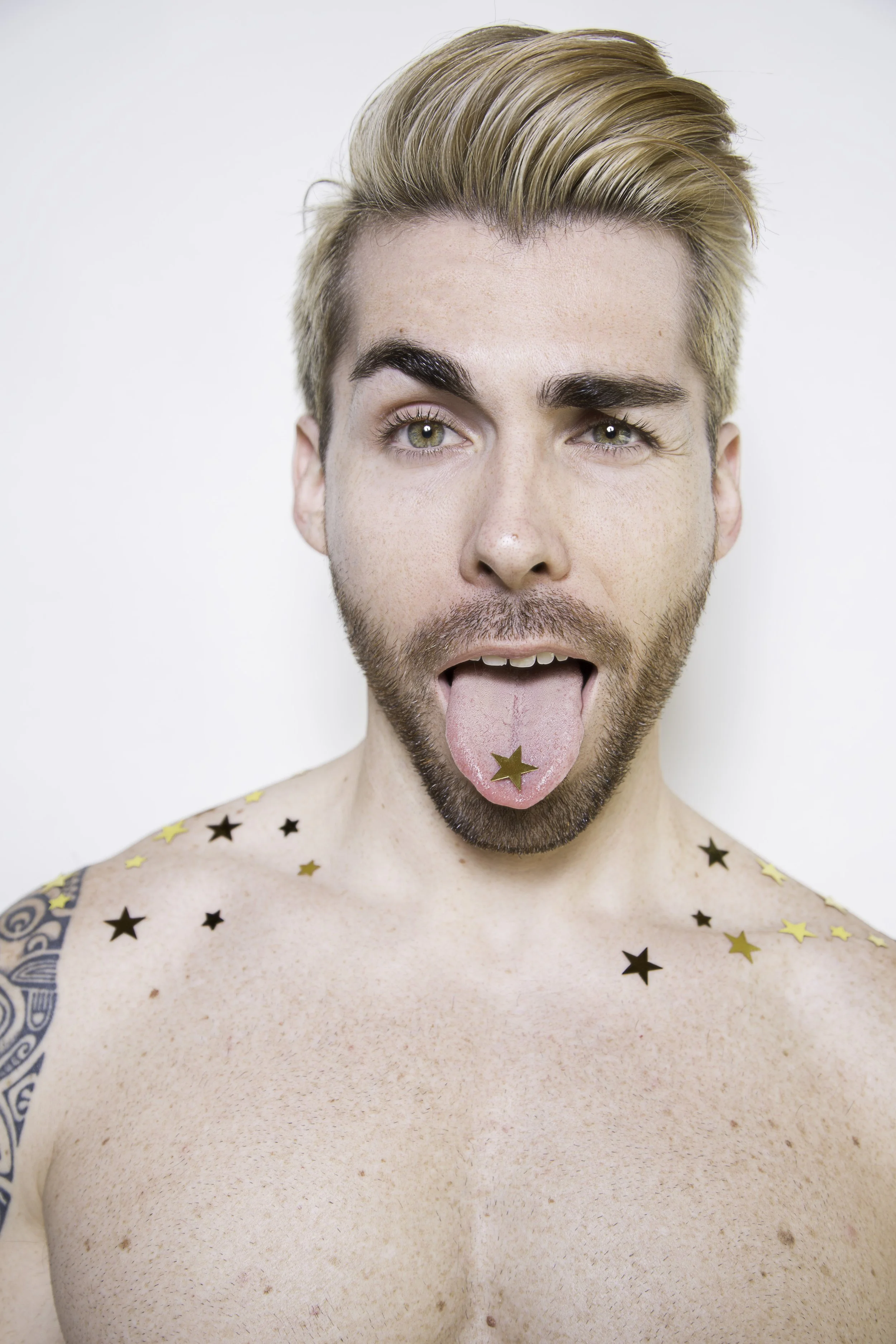 A shirtless young man with styled blond hair, green eyes, and a beard, sticking out his tongue which has a gold star sticker, and gold and black star-shaped confetti on his shoulders and chest, against a plain white background.