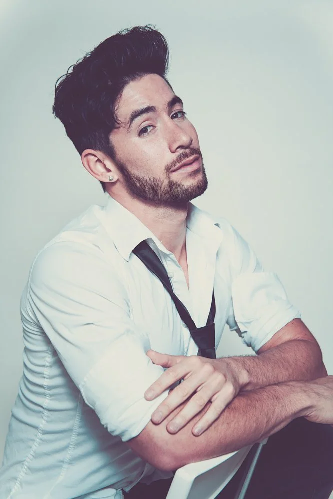 A young man with dark hair, a beard, and earrings, dressed in a white shirt with the sleeves rolled up, sits at a table with his arms crossed and a black tie loosely around his neck, looking wistfully at the camera against a plain light background.