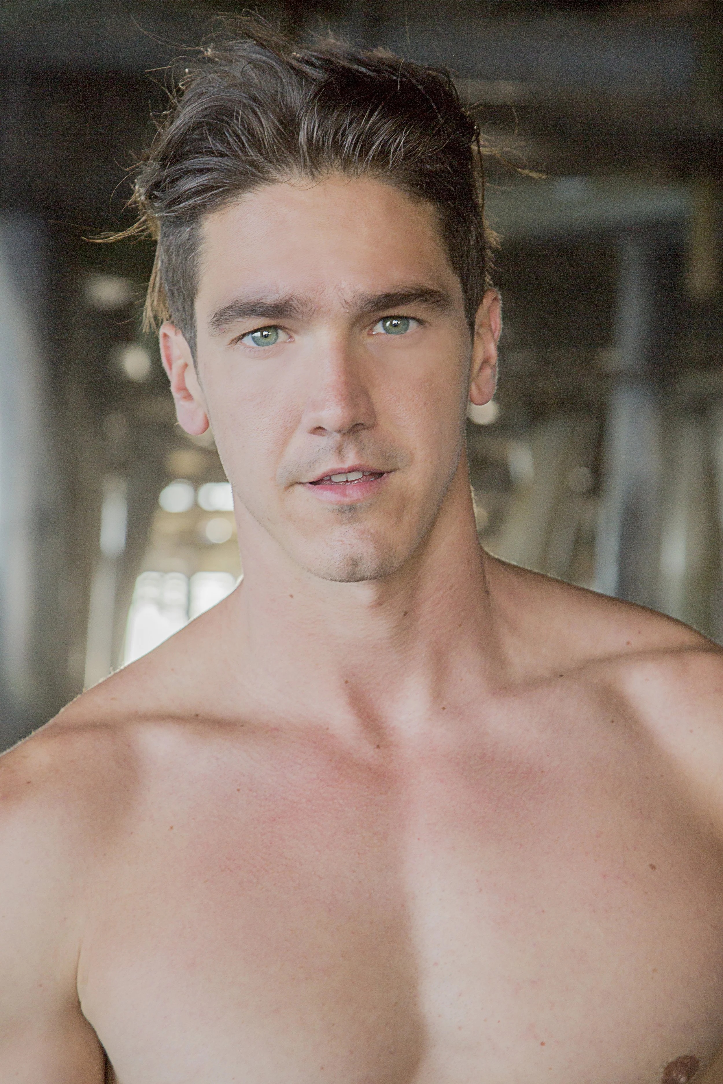 A shirtless young man with blue eyes and dark hair looking directly at the camera, in an industrial setting with metal structures in the background.