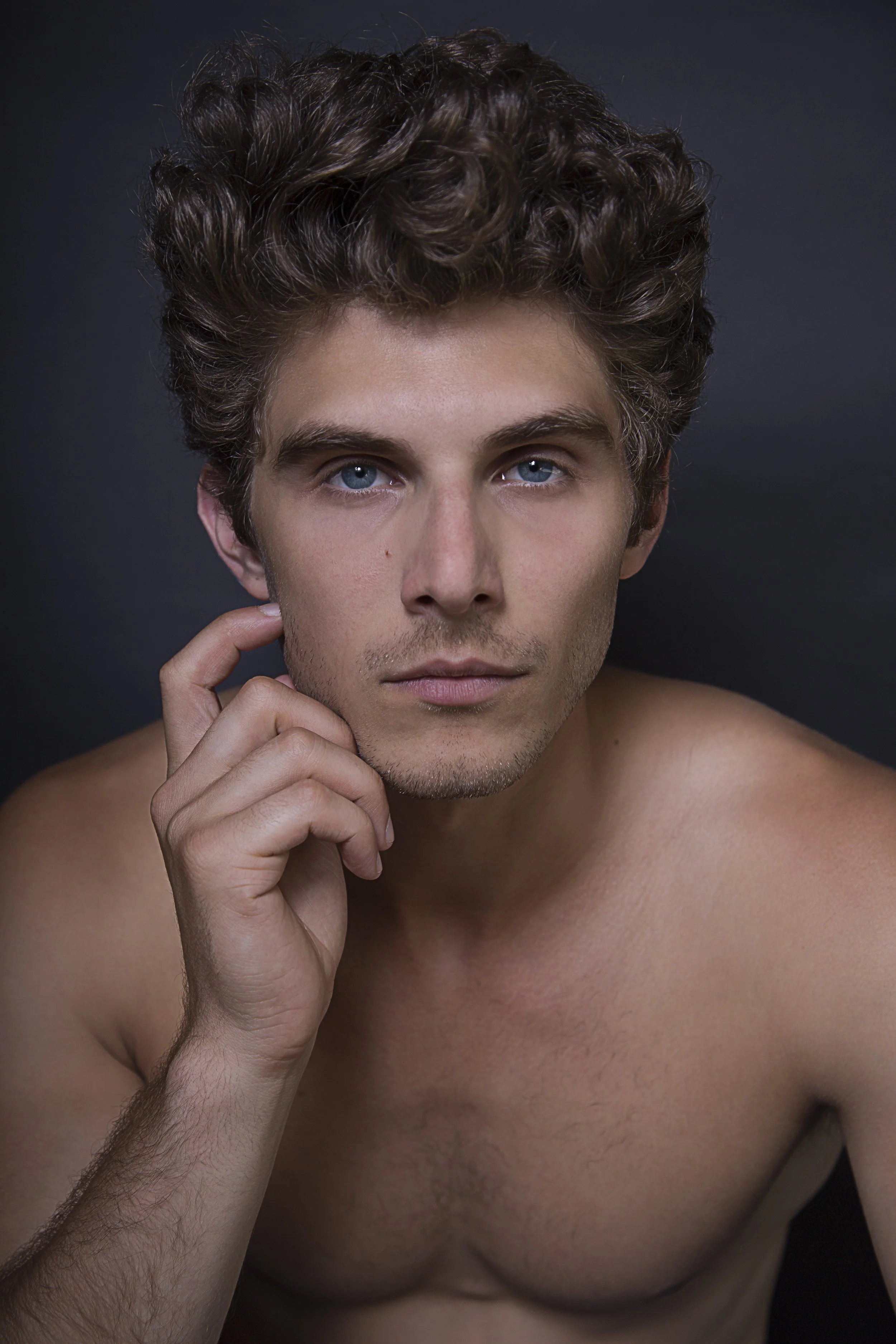 A shirtless young man with curly brown hair and blue eyes, resting his chin on his hand against a dark background.