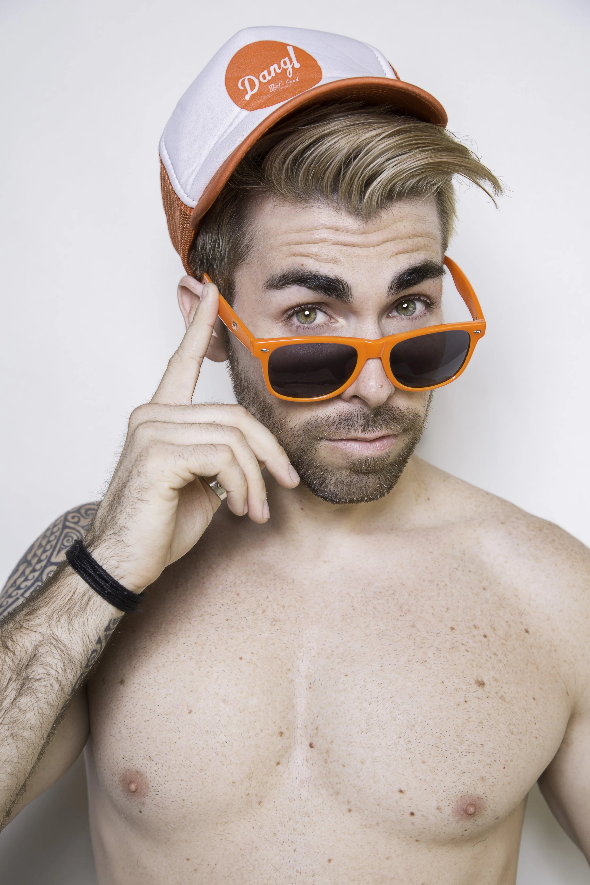 A shirtless young man with sunglasses, a baseball cap, and a tattoo on his arm, pointing to his temple against a plain background.