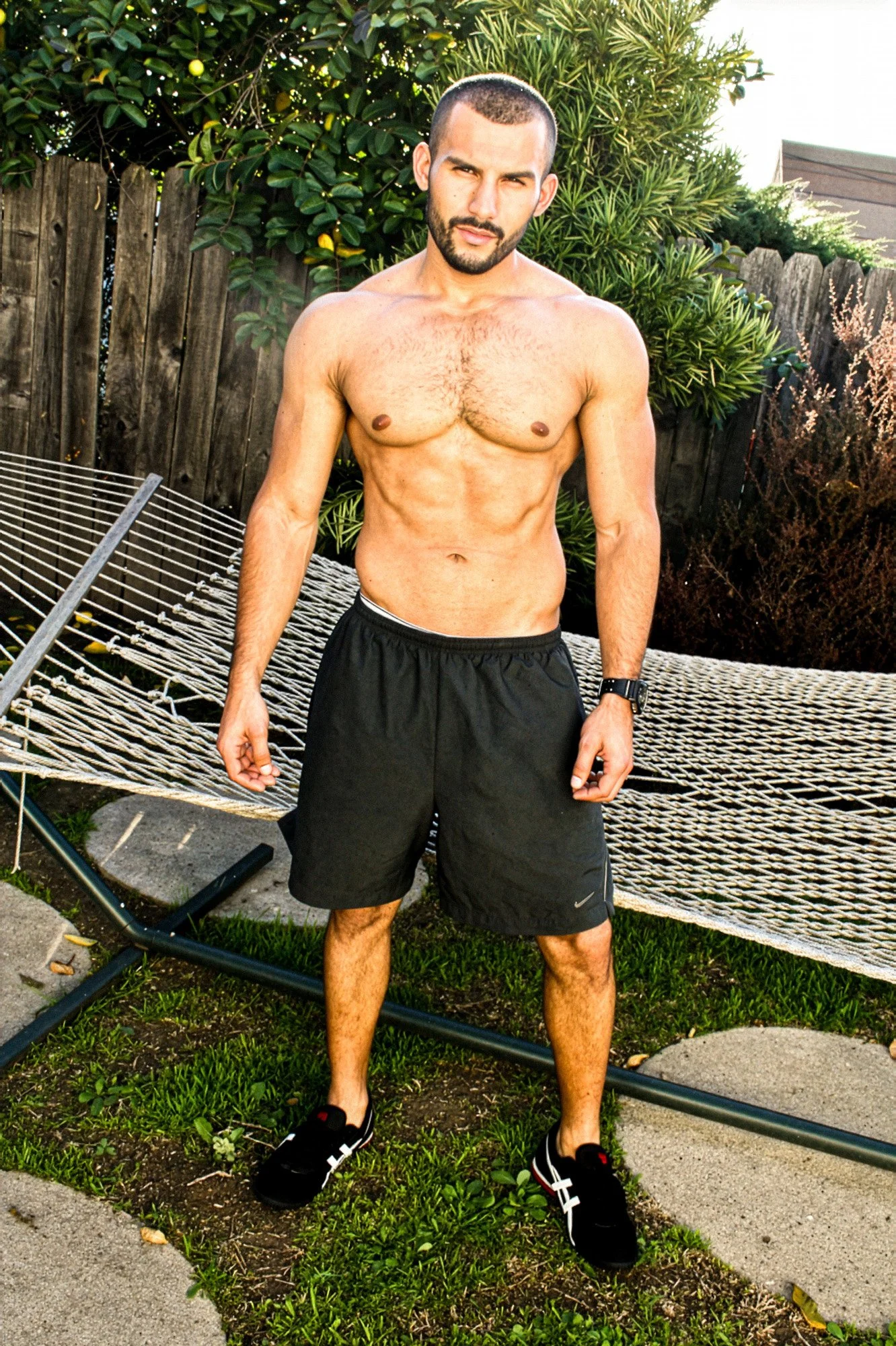 A shirtless man with a beard and short hair standing outdoors next to a hammock in a backyard with plants and a wooden fence.