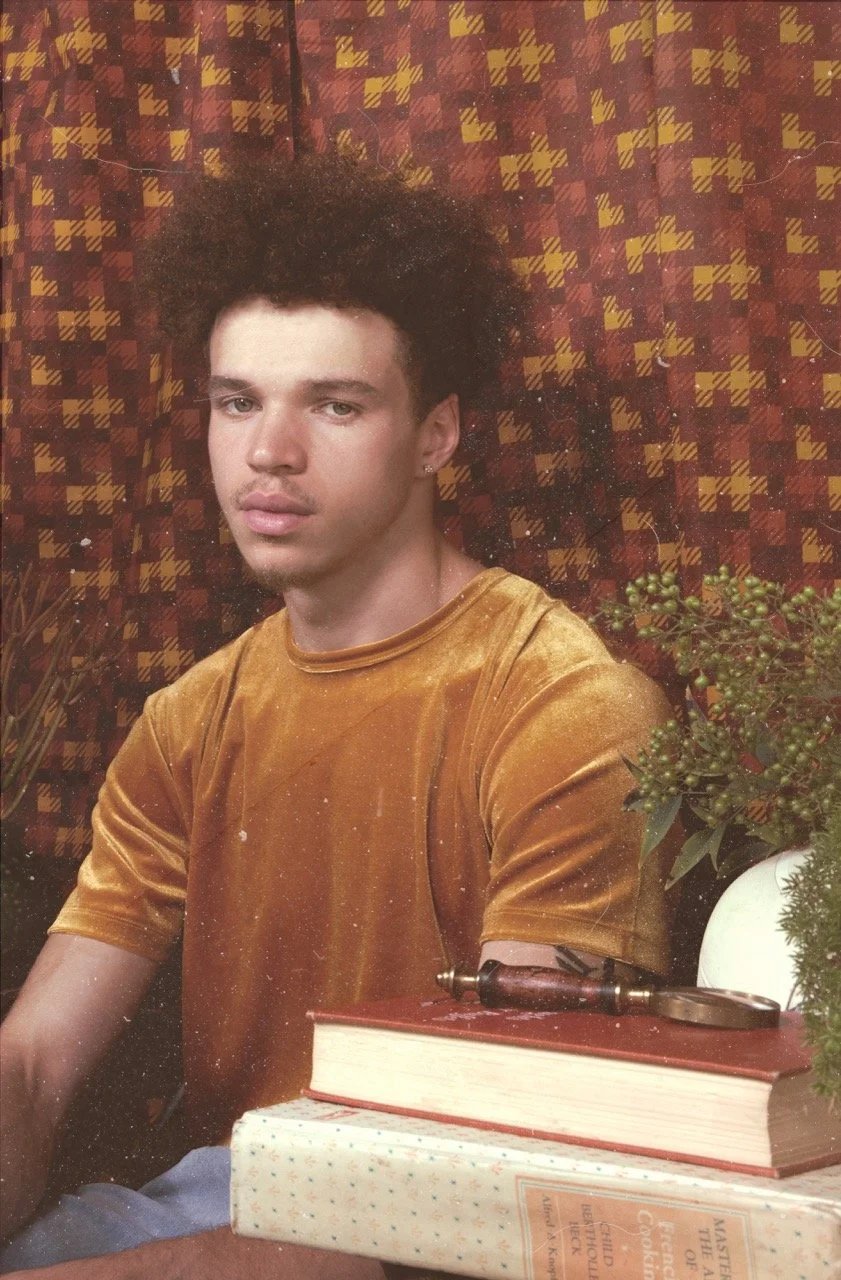 A young man with curly hair and earrings sits indoors in front of a patterned background, wearing a mustard-colored velvet shirt, with books, a small gavel, and a potted plant on the table in front of him.
