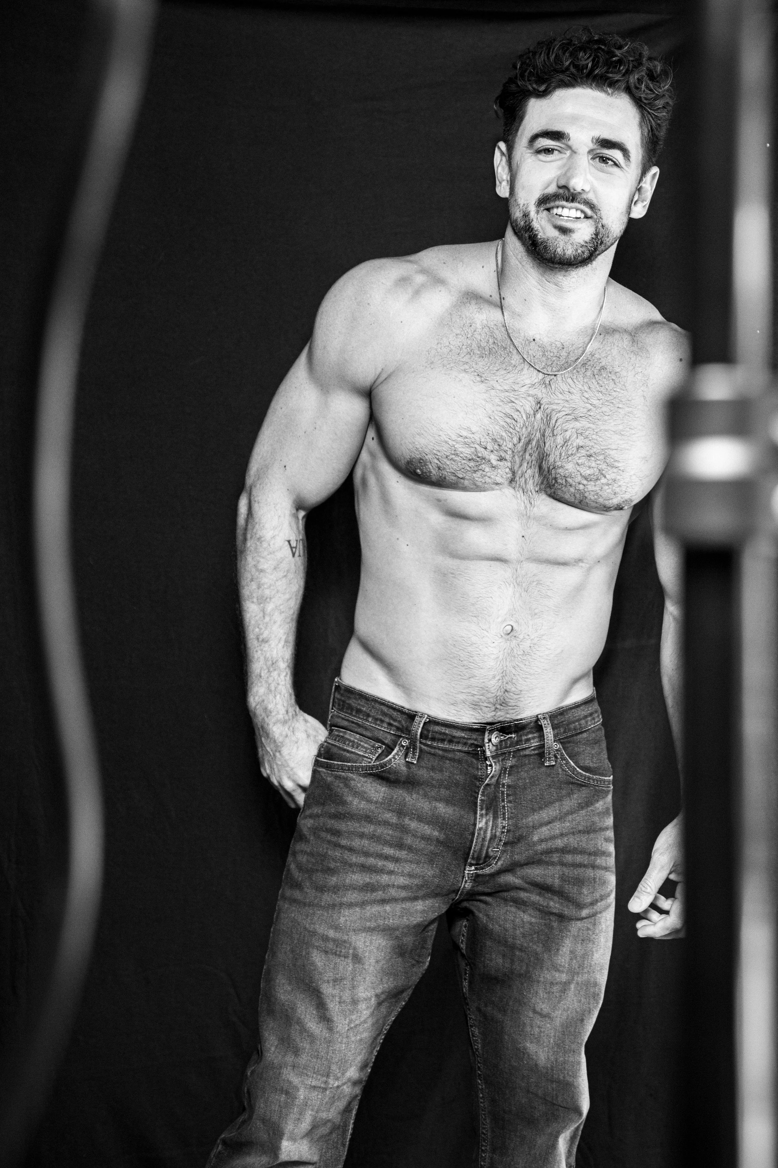 A shirtless man with a beard and curly hair posing in front of a dark background, wearing jeans and a necklace.