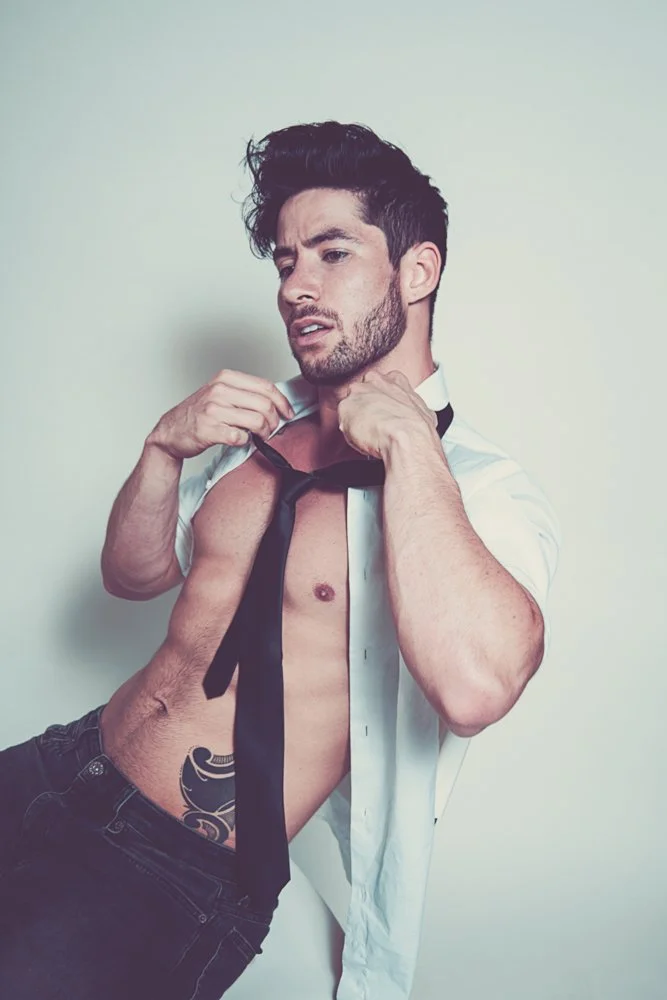 A shirtless man with dark hair and a beard, wearing a white shirt and a loosened black tie, holds the collar of his shirt as he leans against a plain white wall.