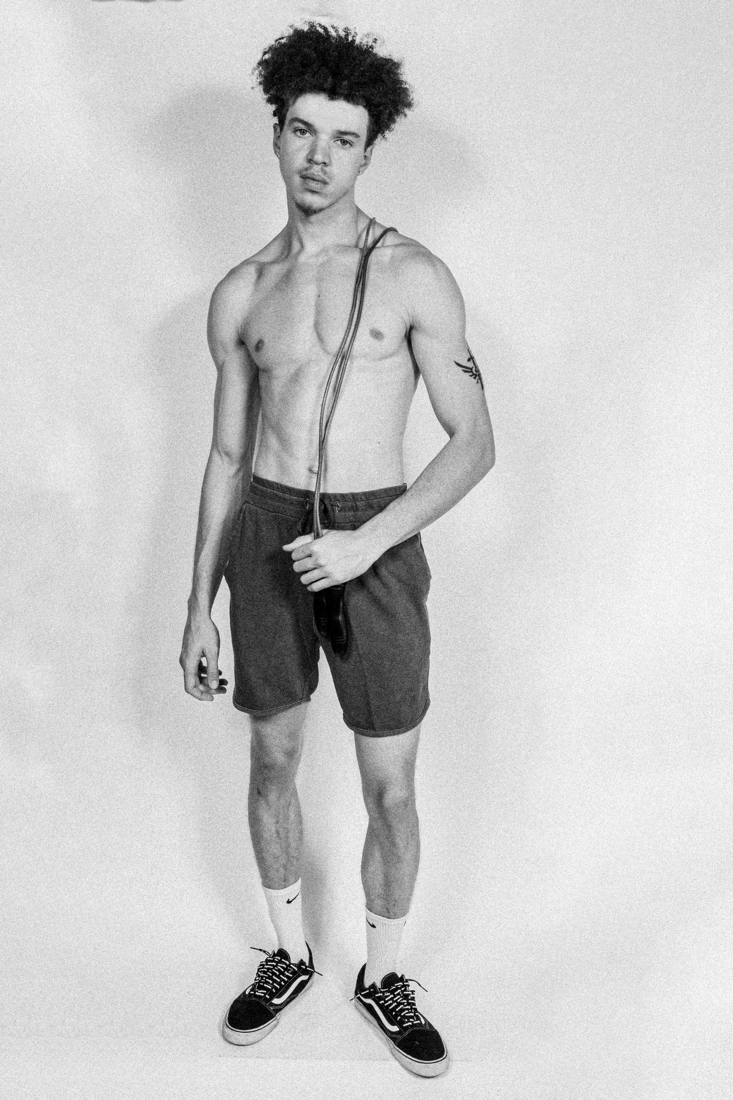 Black and white photo of a shirtless young man with curly hair, wearing shorts, Nike socks, and sneakers, holding a jump rope, standing against a plain background.