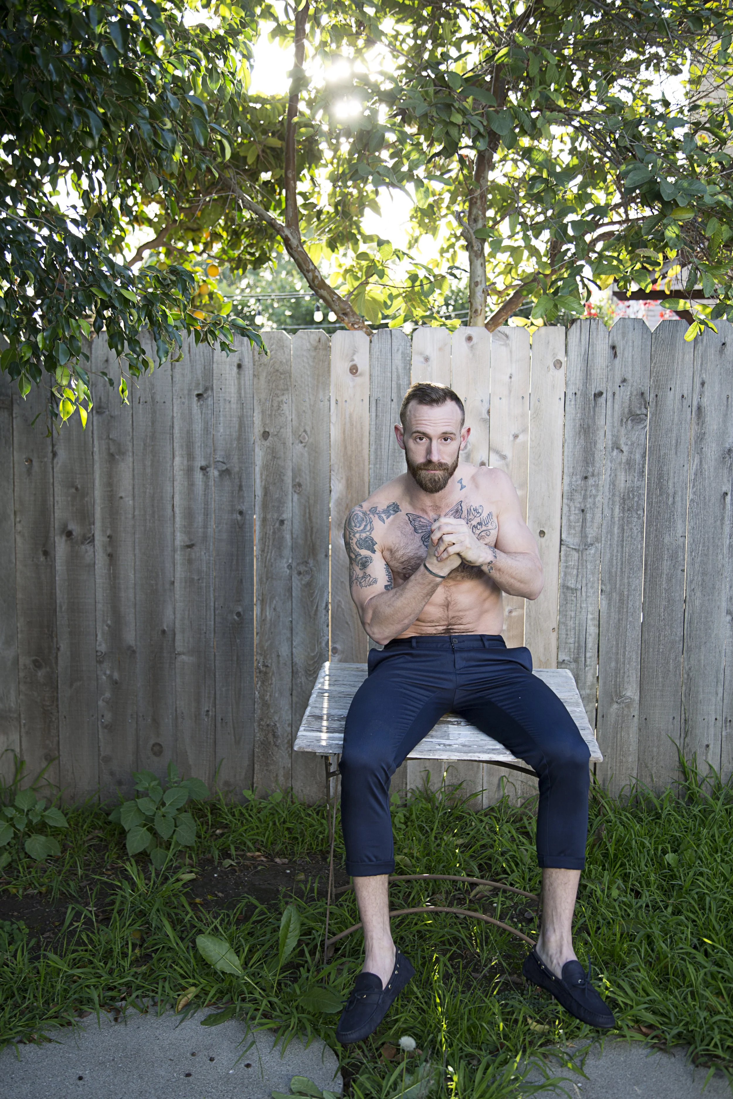 A shirtless man with tattoos sits on a wooden bench outdoors, with a wooden fence behind him and a tree with green leaves overhead. The sun is shining through the leaves, creating a bright effect.