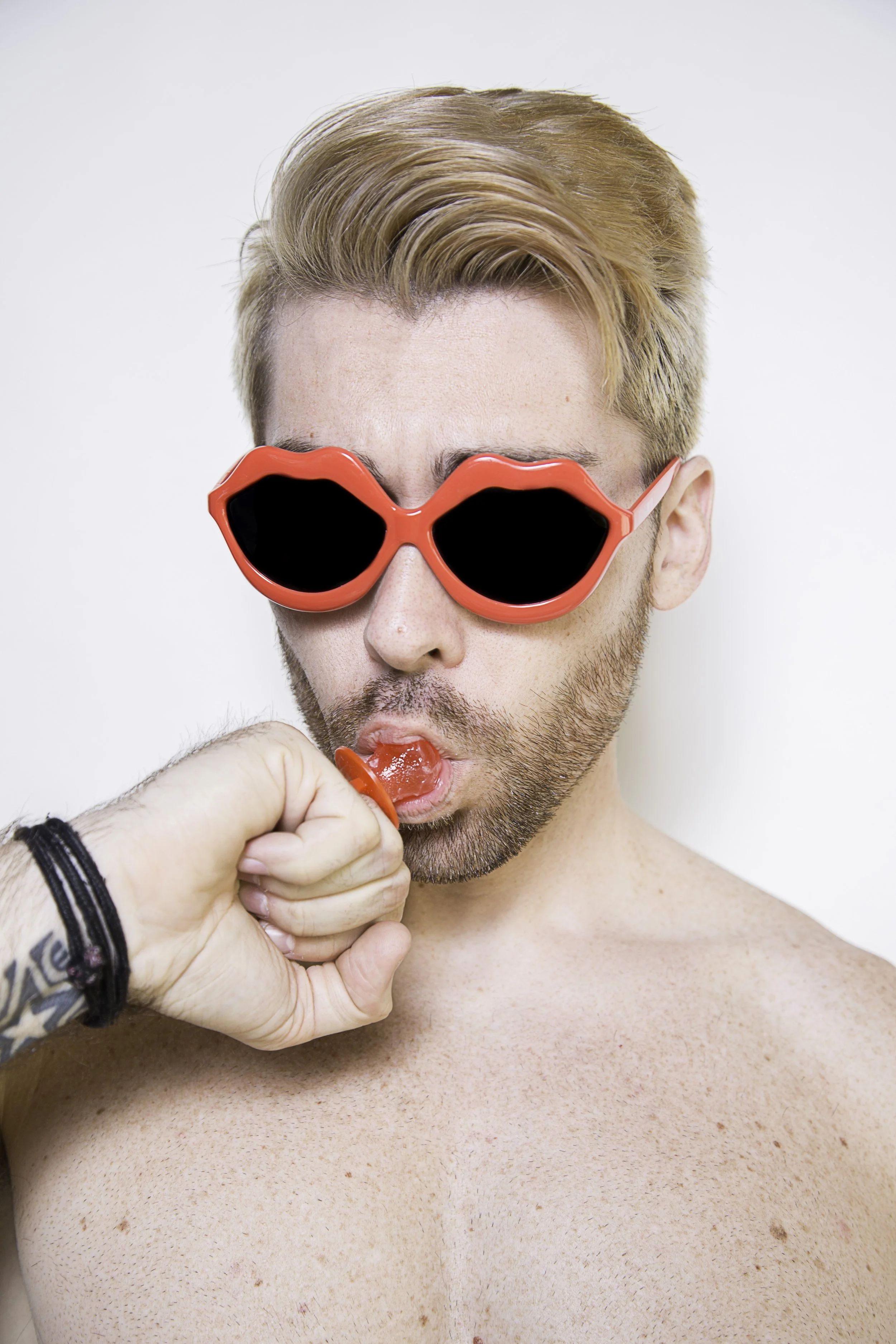 A shirtless man with blond hair and sunglasses shaped like red lips is licking a red popsicle being held by another person with a tattooed wrist.