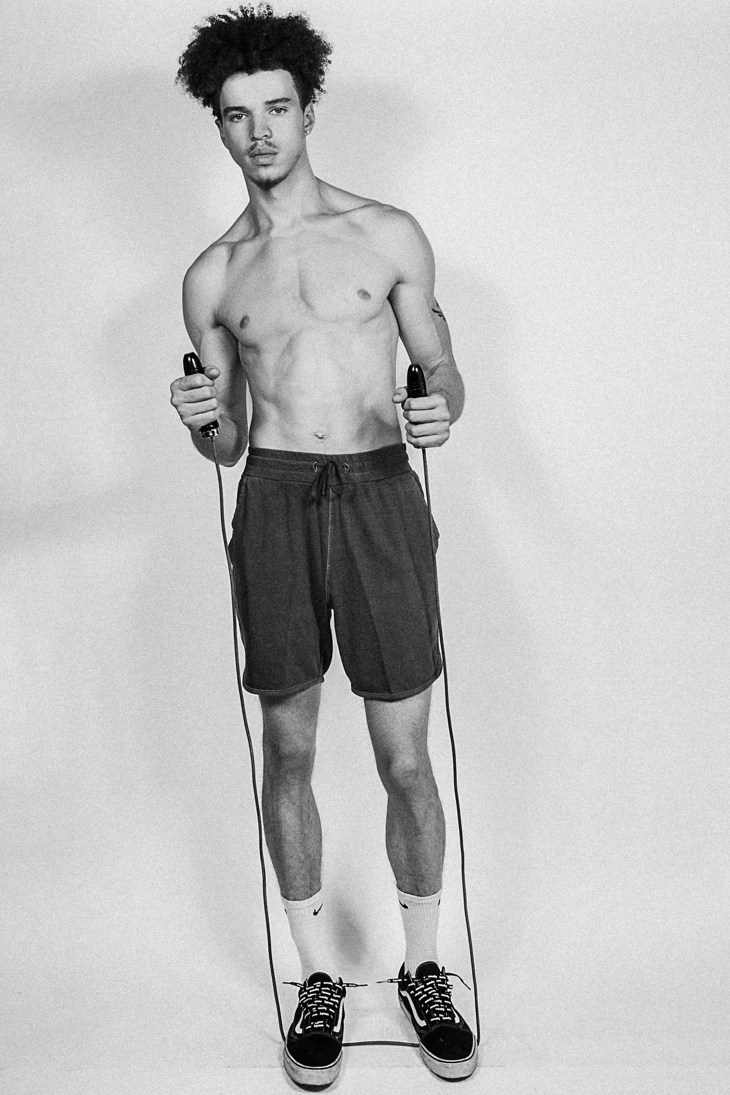 A shirtless young man with curly hair, wearing shorts, sneakers, and Nike socks, holding jump ropes in both hands, standing against a plain background.