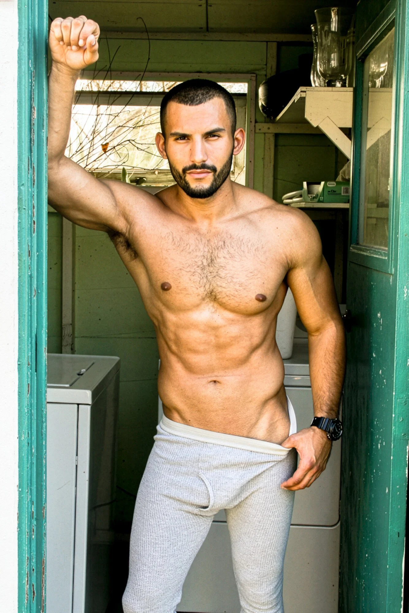 A shirtless man with a muscular build standing inside a small green wooden structure, raising one arm and holding his waistband with the other, looking into the camera.