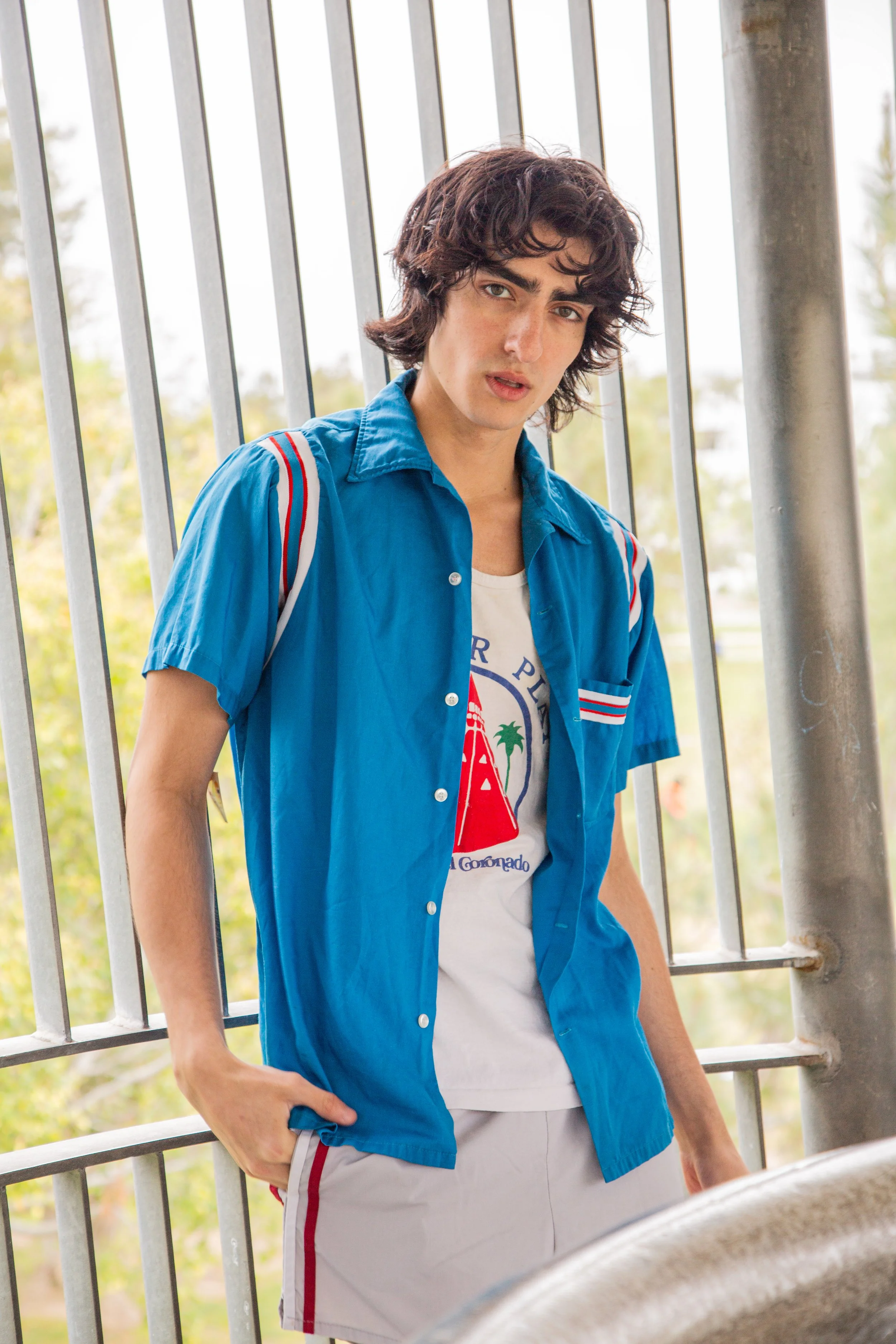 A young man with dark wavy hair wearing a blue short-sleeved shirt over a white graphic t-shirt and gray shorts, standing next to a metal fence outdoors, looking at the camera.