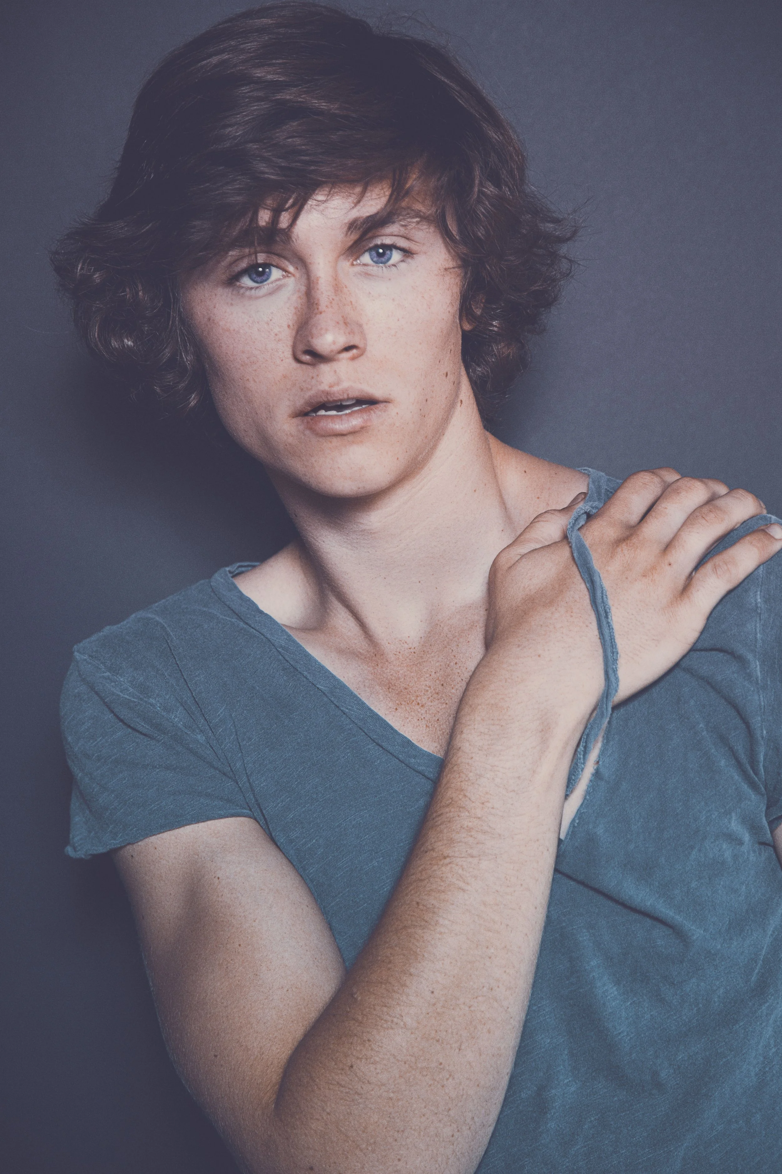 A person with short, curly brown hair and blue eyes wearing a blue shirt and touching their shoulder.