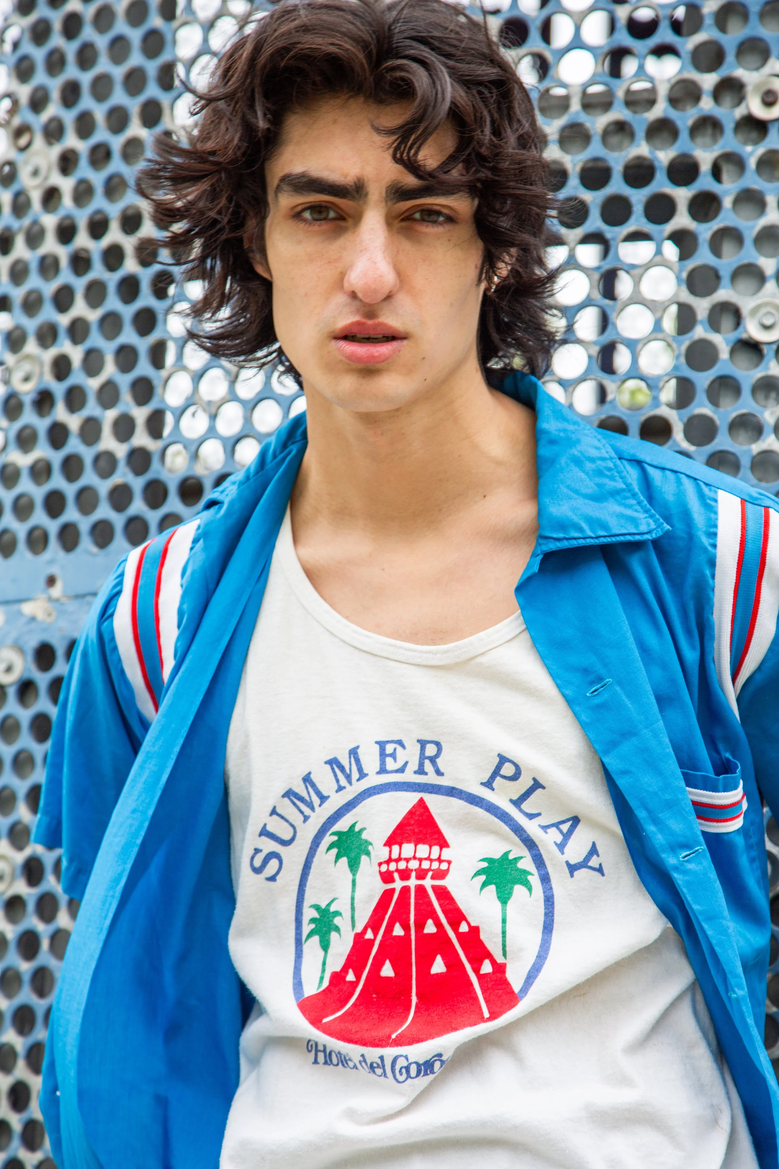 A young man with curly dark hair and light skin wearing a white T-shirt with a colorful graphic and text, and an unbuttoned blue jacket, standing against a perforated metal background.
