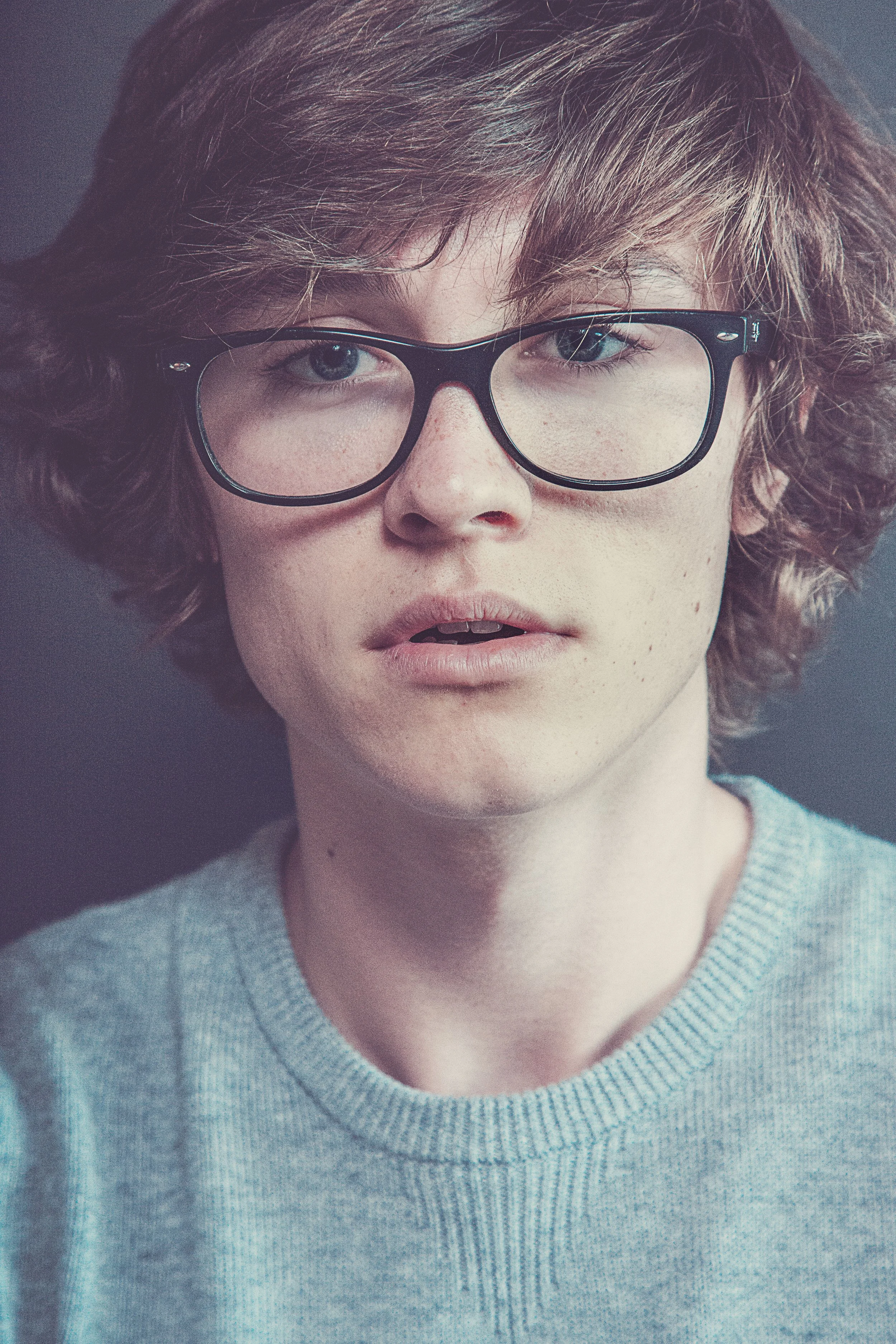 Close-up of a young person with curly brown hair, wearing large black glasses and a light blue sweater, with a neutral facial expression.