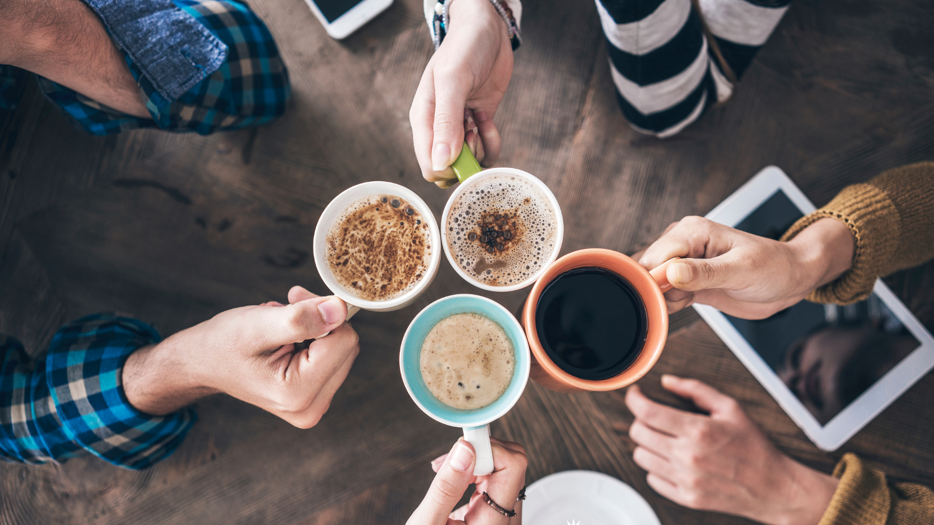 Four people holding colorful coffee mugs, clinking them together over a wooden table. There are some digital devices and a cup of coffee on the table.