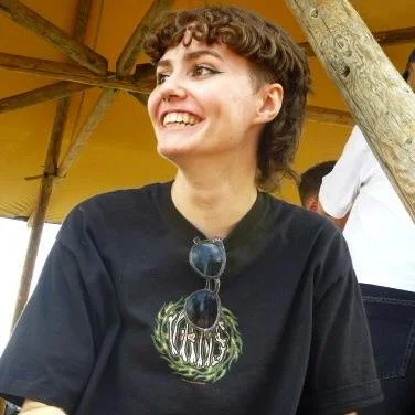 A smiling woman (CJ Harris) with short curly hair wearing a black T-shirt and sunglasses hanging from her shirt, sitting outdoors under a yellow canopy.