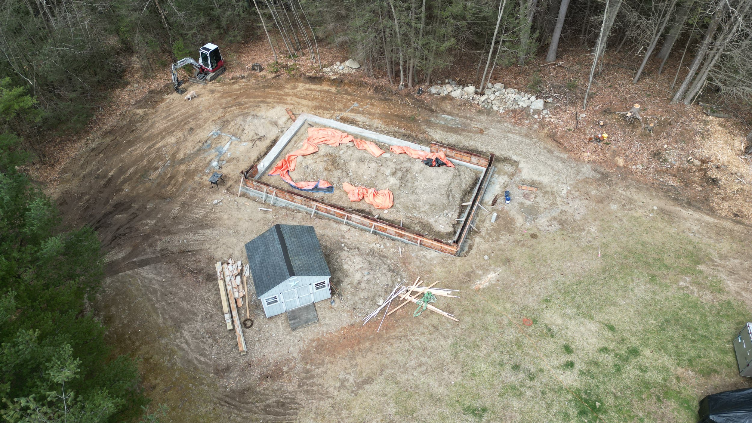 An aerial view of a construction site with a foundation being built, a small shed, a pile of wooden planks, and machinery in the background surrounded by trees.
