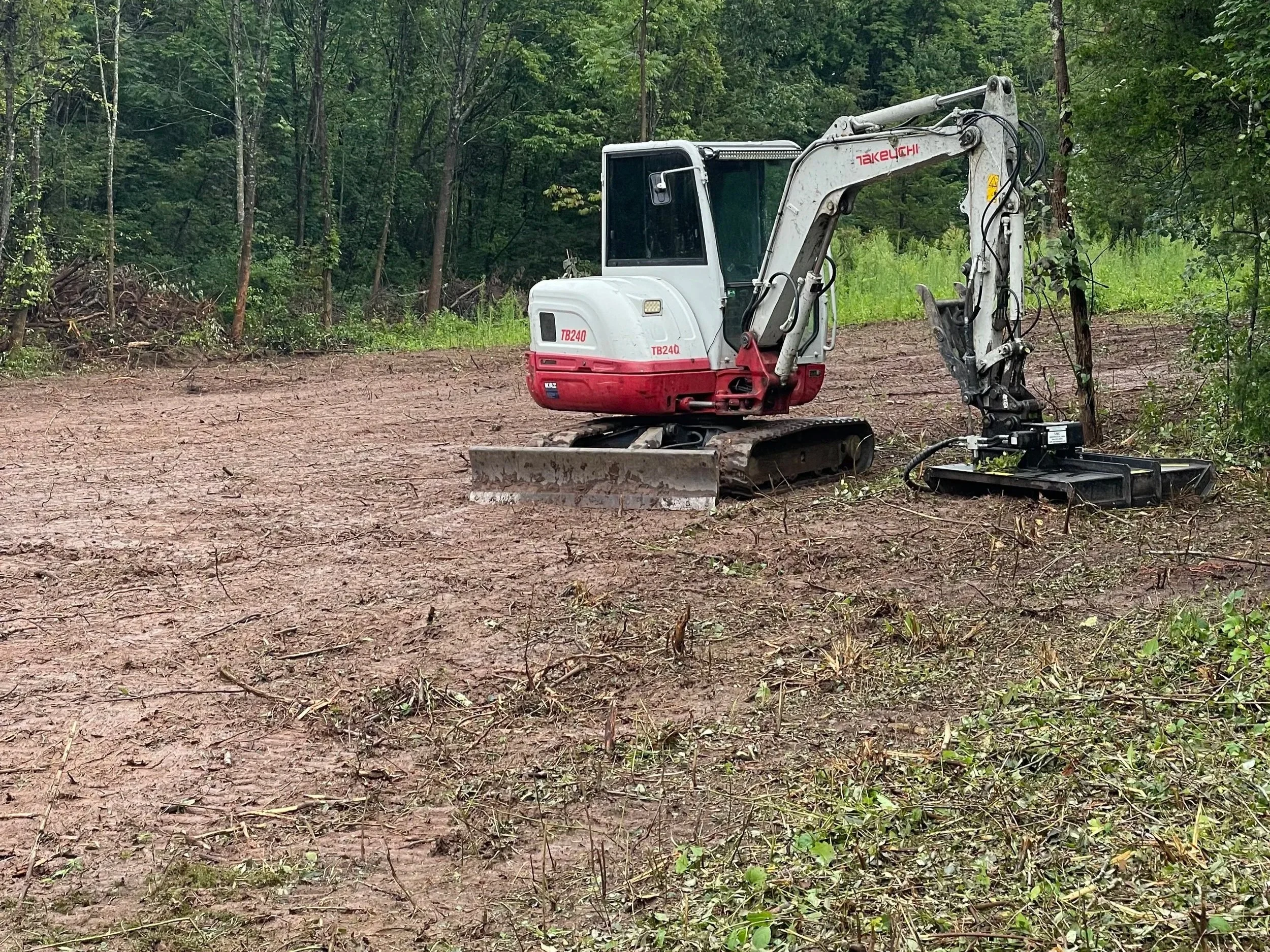 A small white and red excavator on a cleared patch of land in a forested area.