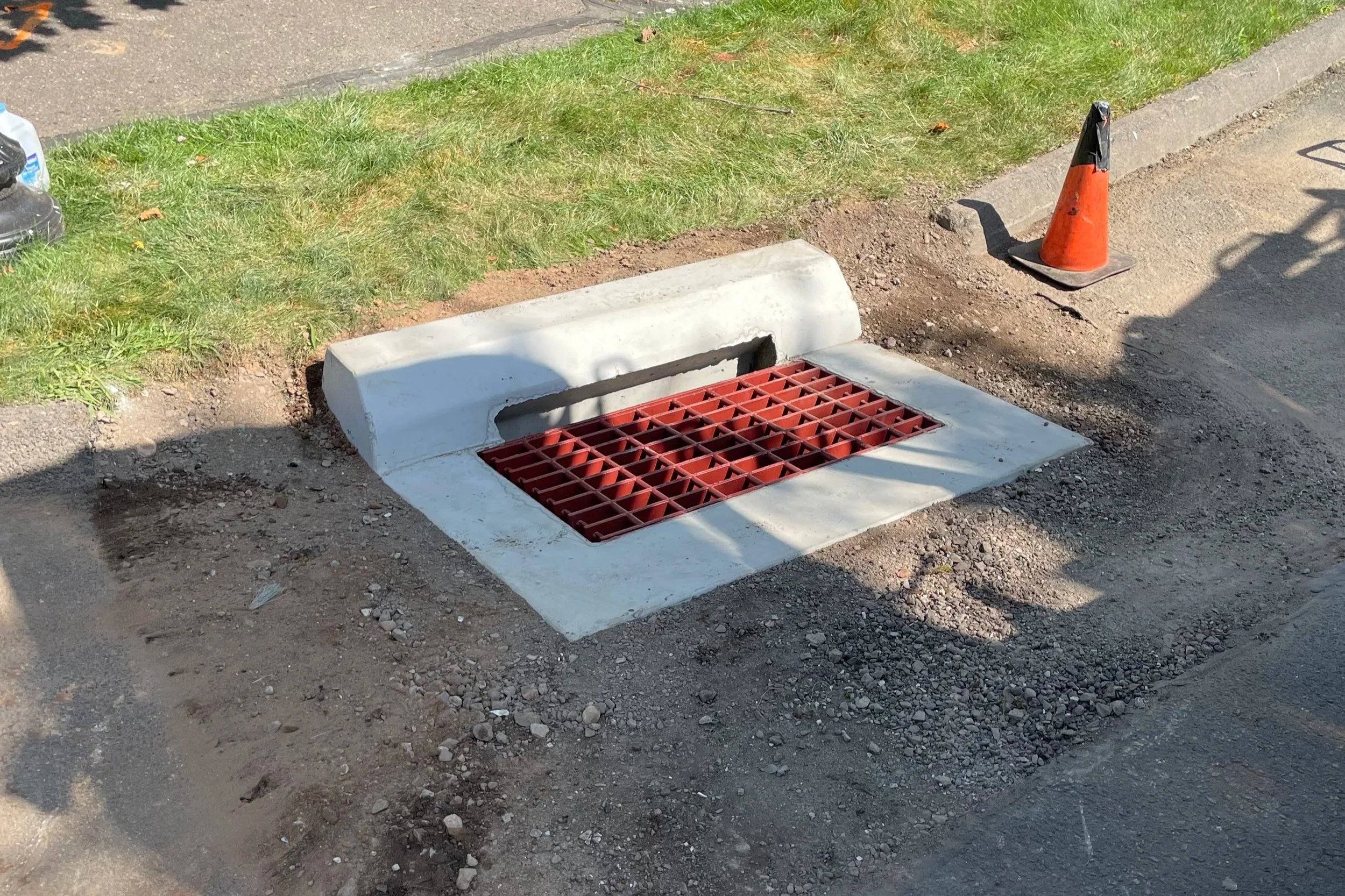 A street drain with a red grate surrounded by a concrete border, partially covered with fresh white paint, located on the side of a street next to a grassy area and an orange traffic cone.