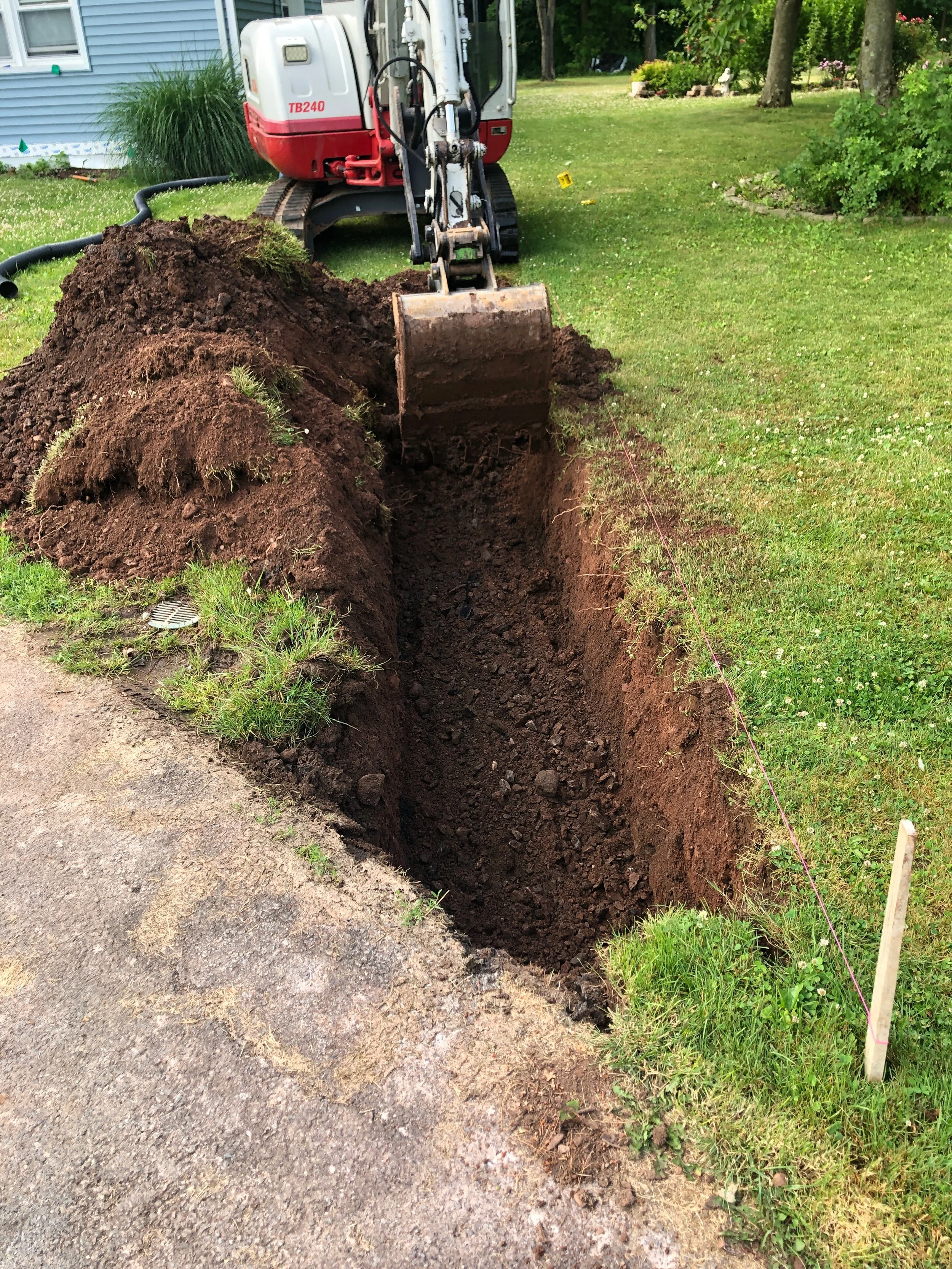 A small excavator digging a trench in a grassy yard next to a house, with a string line marking the trench's boundary.