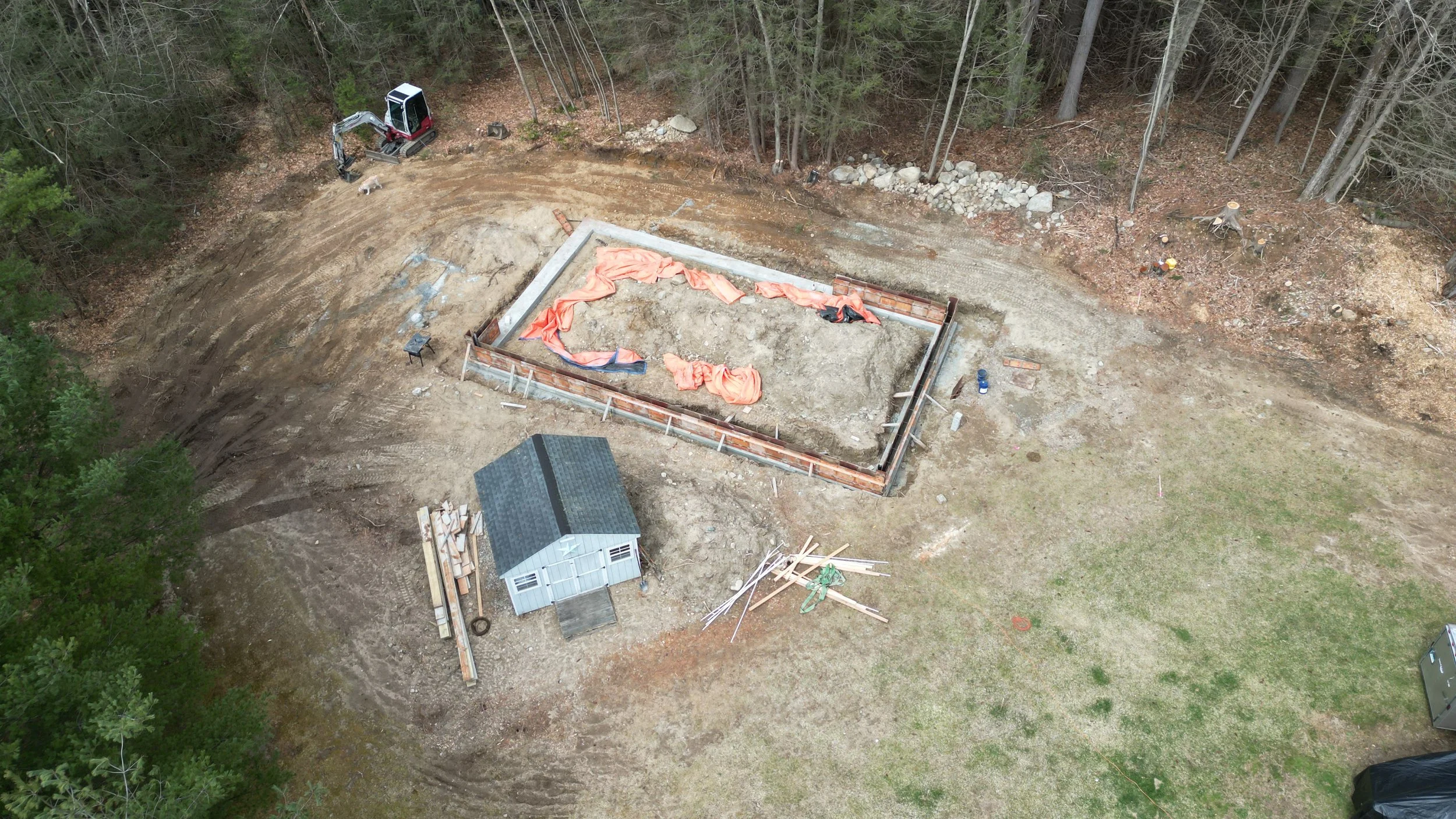 Aerial view of a construction site with a foundation outline, a small shed, and construction tools, set in a cleared area near a wooded forest.