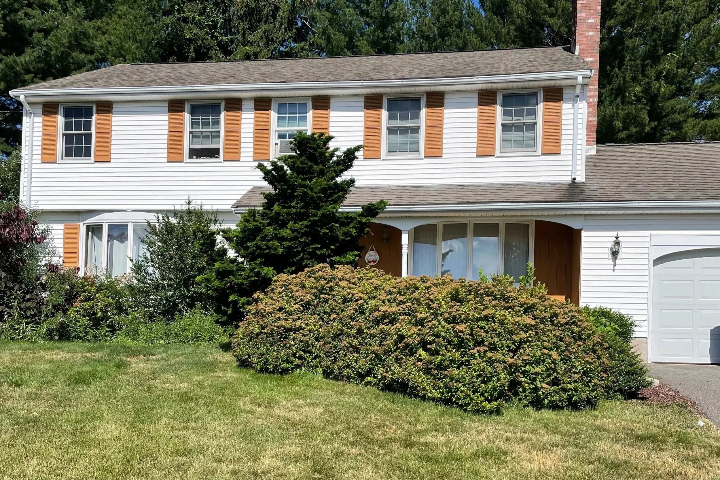 Front view of a two-story suburban house with white siding, brown shutters, a bay window, a front door, a garage, and landscaped front yard with bushes and grass.