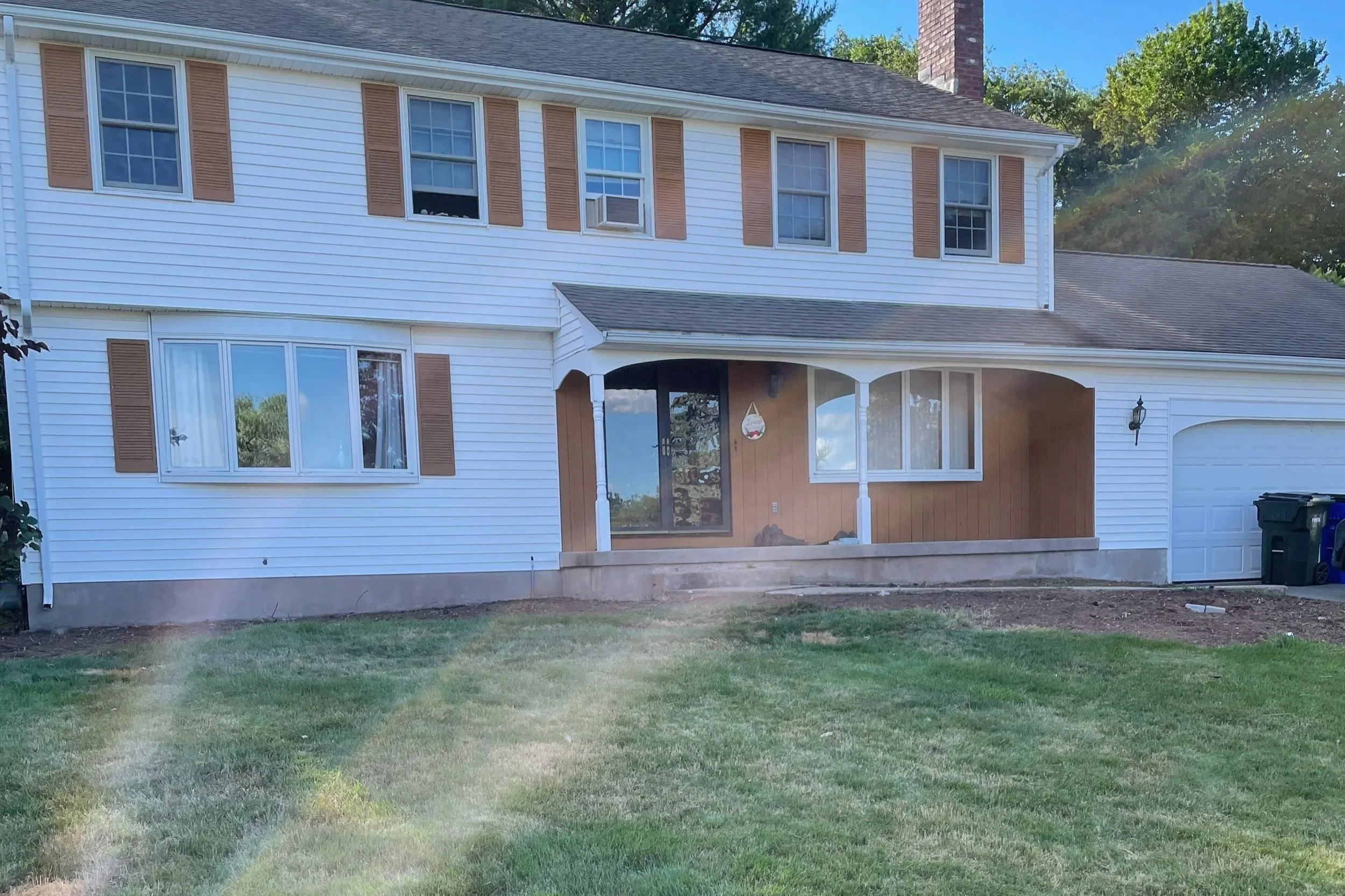 A two-story house with white siding, brown shutters, and a small porch with steps. The front yard has a grassy lawn and is partly shaded. There are trash bins near the garage, and the house has a chimney and multiple windows.