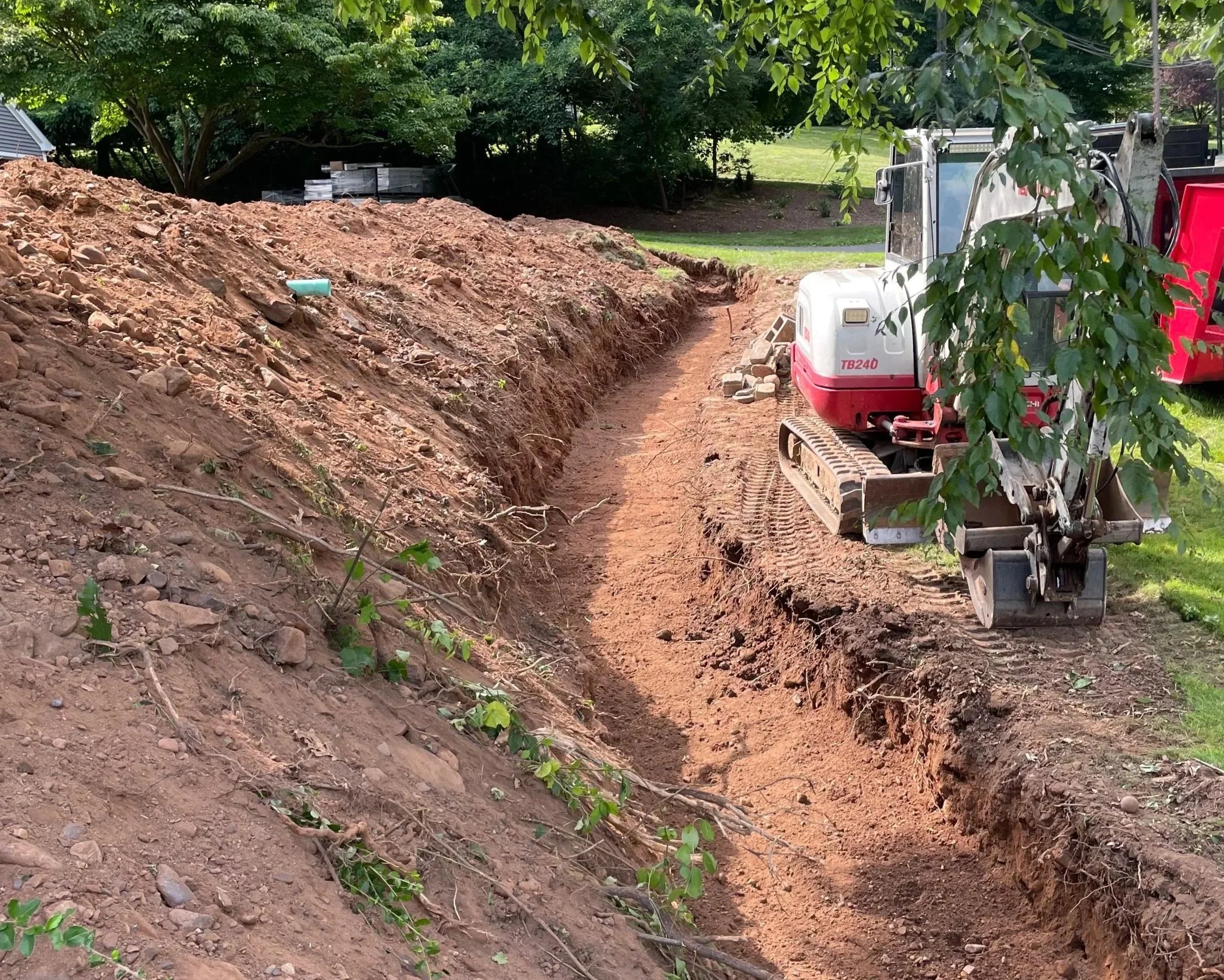 A small excavator working on a dirt trench in a residential yard with green trees and grass in the background.