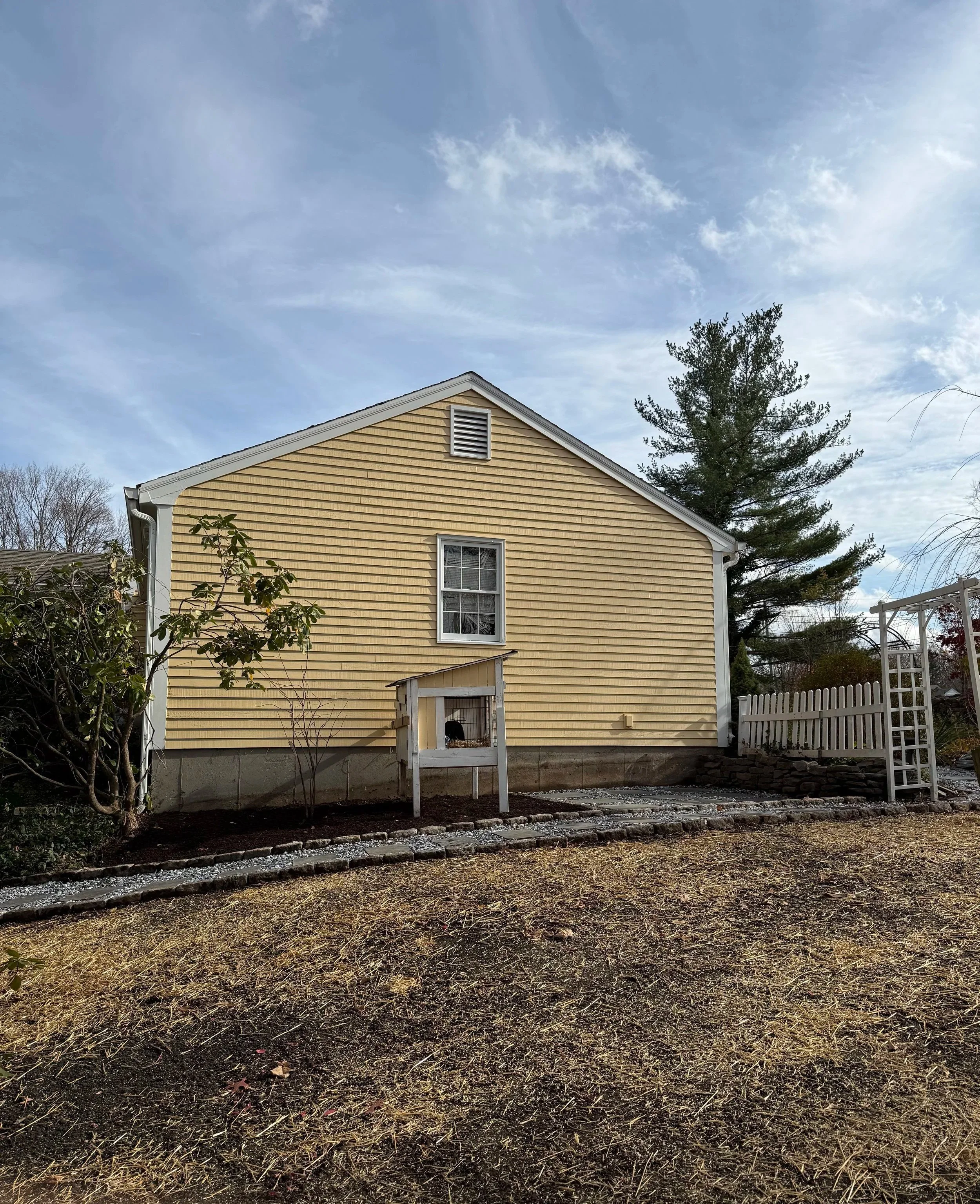 Rear view of a yellow house with white trim, a small window, a weathered small shed, a white picket fence, and trees, under a partly cloudy sky.