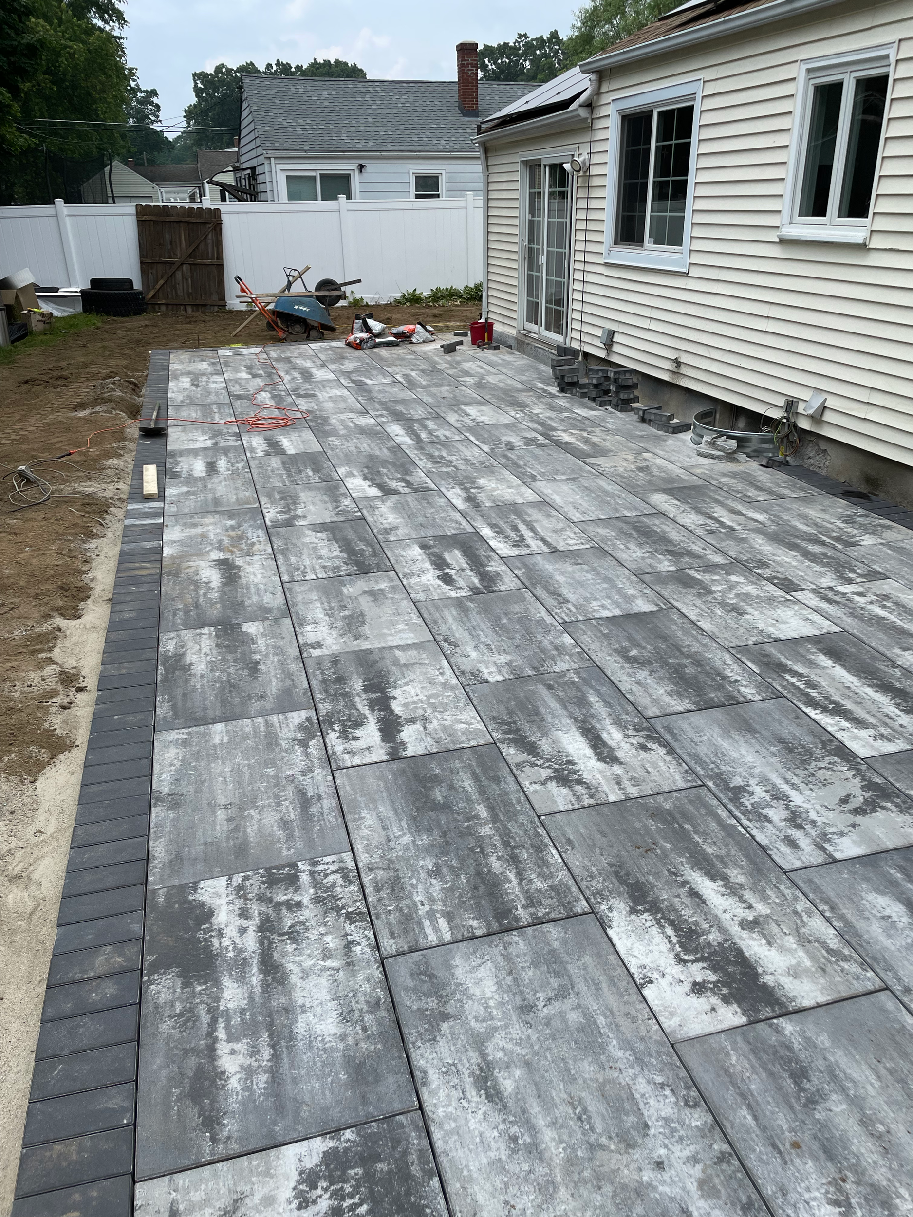 Under construction backyard patio with large gray tiles, shown with some construction tools and materials, adjacent to a house with white siding and a sliding glass door.