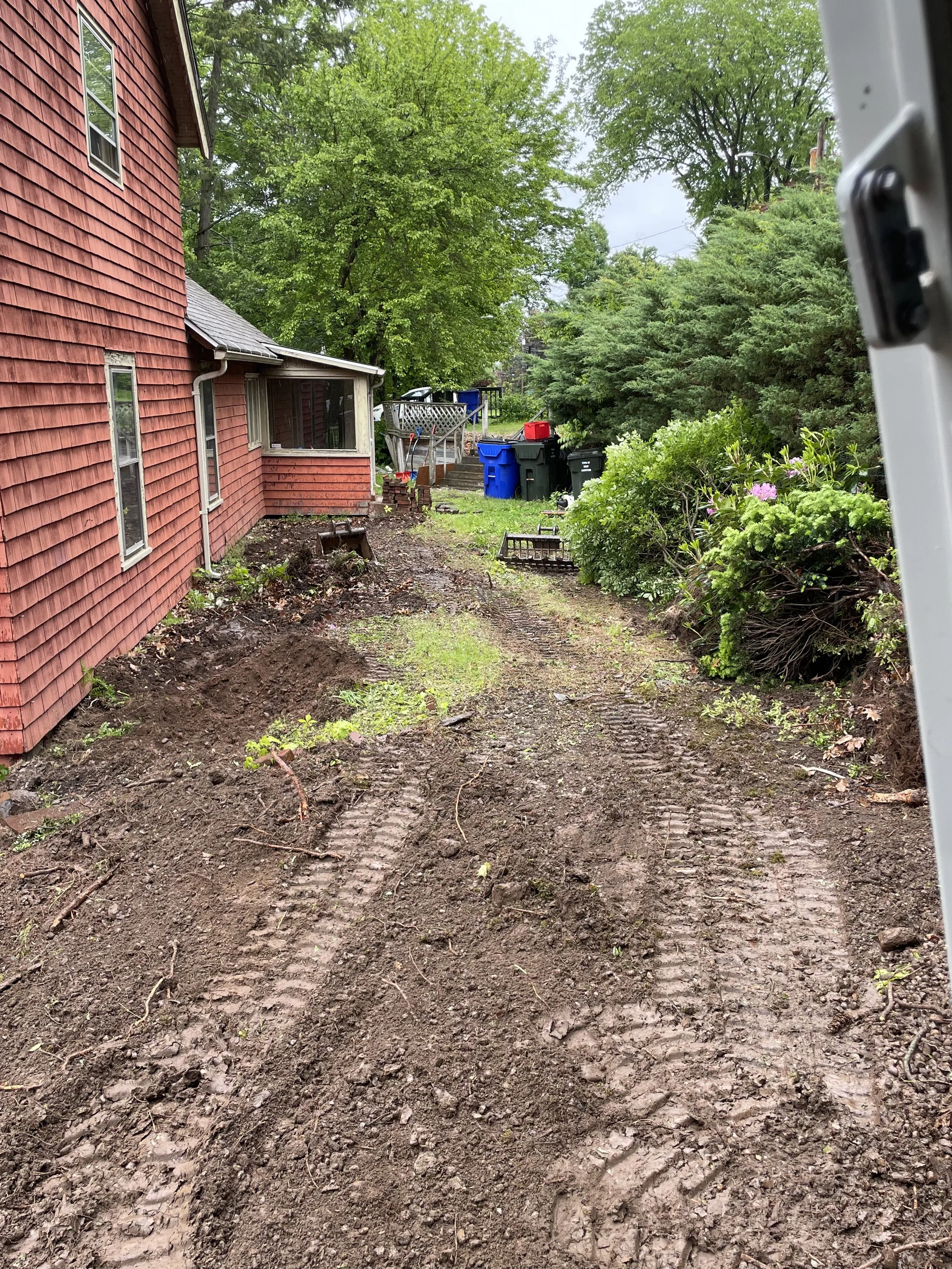 A dirt backyard with tire tracks and patches of grass, next to a red house with three windows, surrounded by green trees and bushes, with trash bins and a wooden fence in the background.