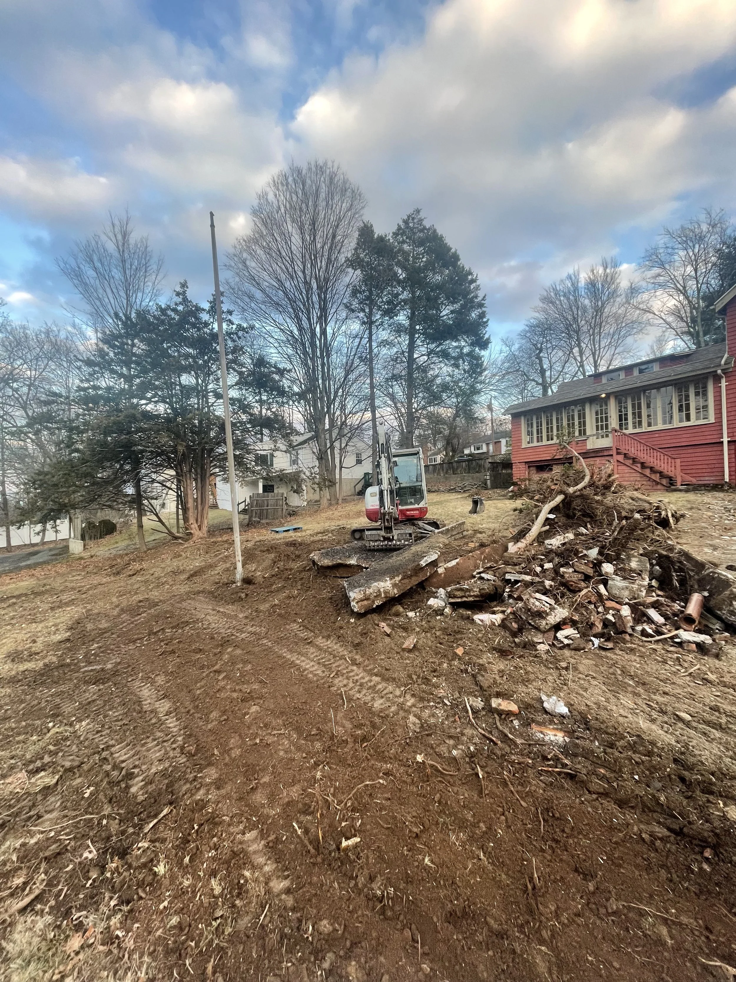 A construction site with a small excavator on a dirt area, debris like broken brick and wood nearby, and residential houses and trees in the background under a partly cloudy sky.