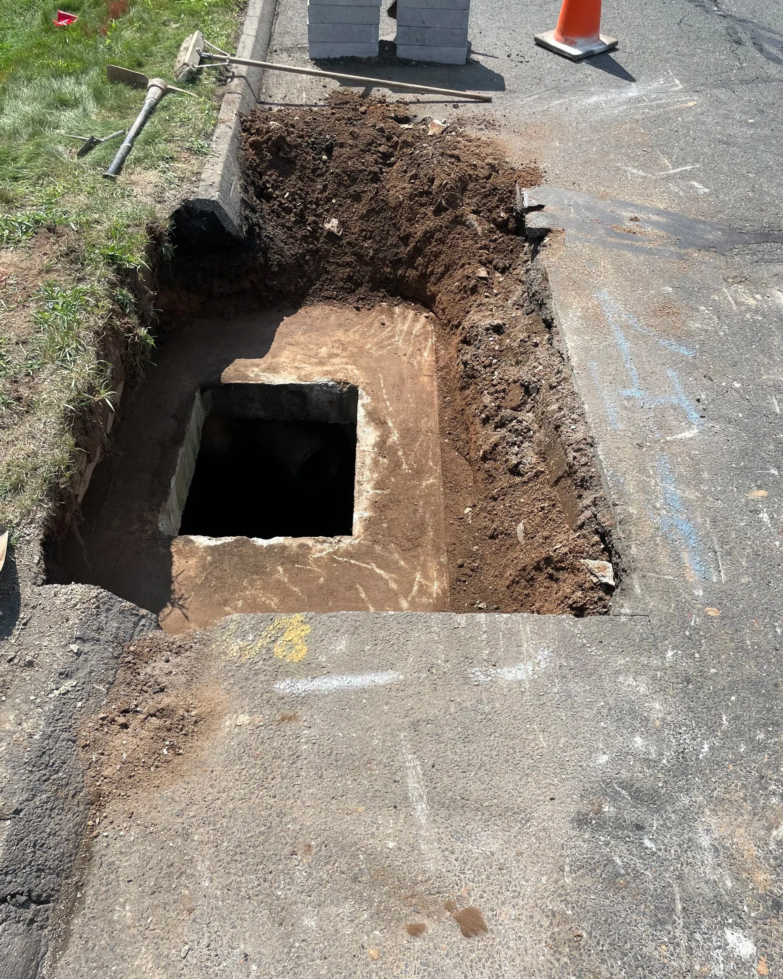 Open excavation in the sidewalk showing underground utility or sewer access with a square opening and surrounding asphalt and dirt