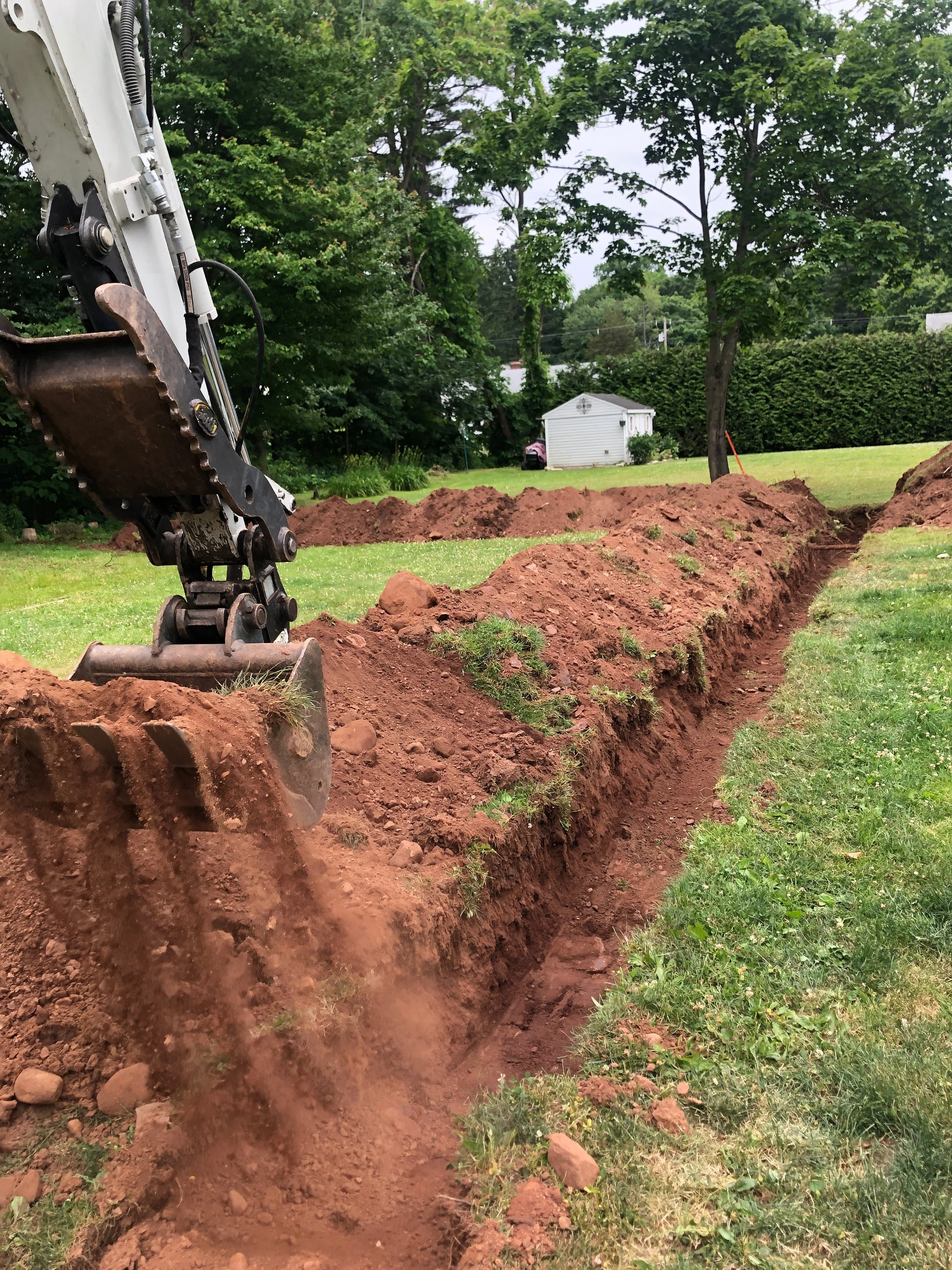 Excavation of a trench in a grassy yard with a backhoe digger. The trench runs across the yard, and the soil is piled along its edges. In the background, there are trees, a small shed, and a lawn.