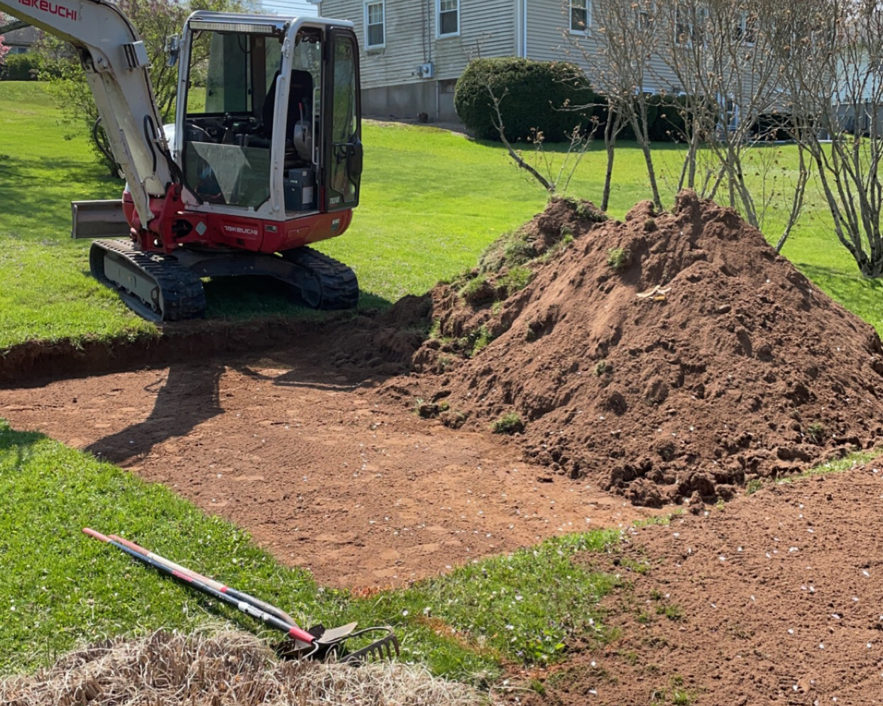 A mini excavator digging a hole in a backyard with a mound of dirt next to it, surrounded by grass and a house in the background.