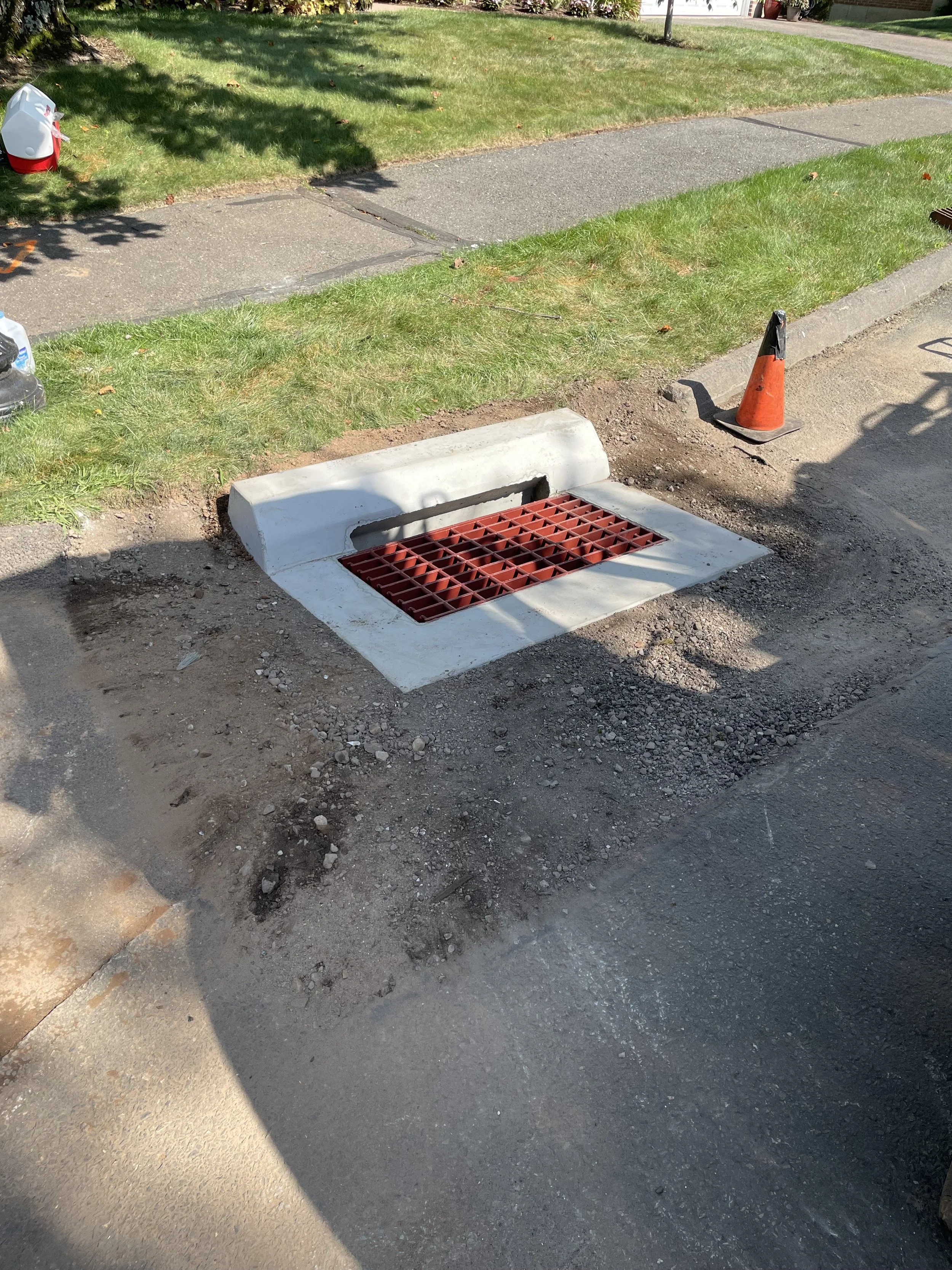 Street construction site with a drain in the road, a traffic cone, and a shadow of a person, in a residential neighborhood with grass and sidewalk.