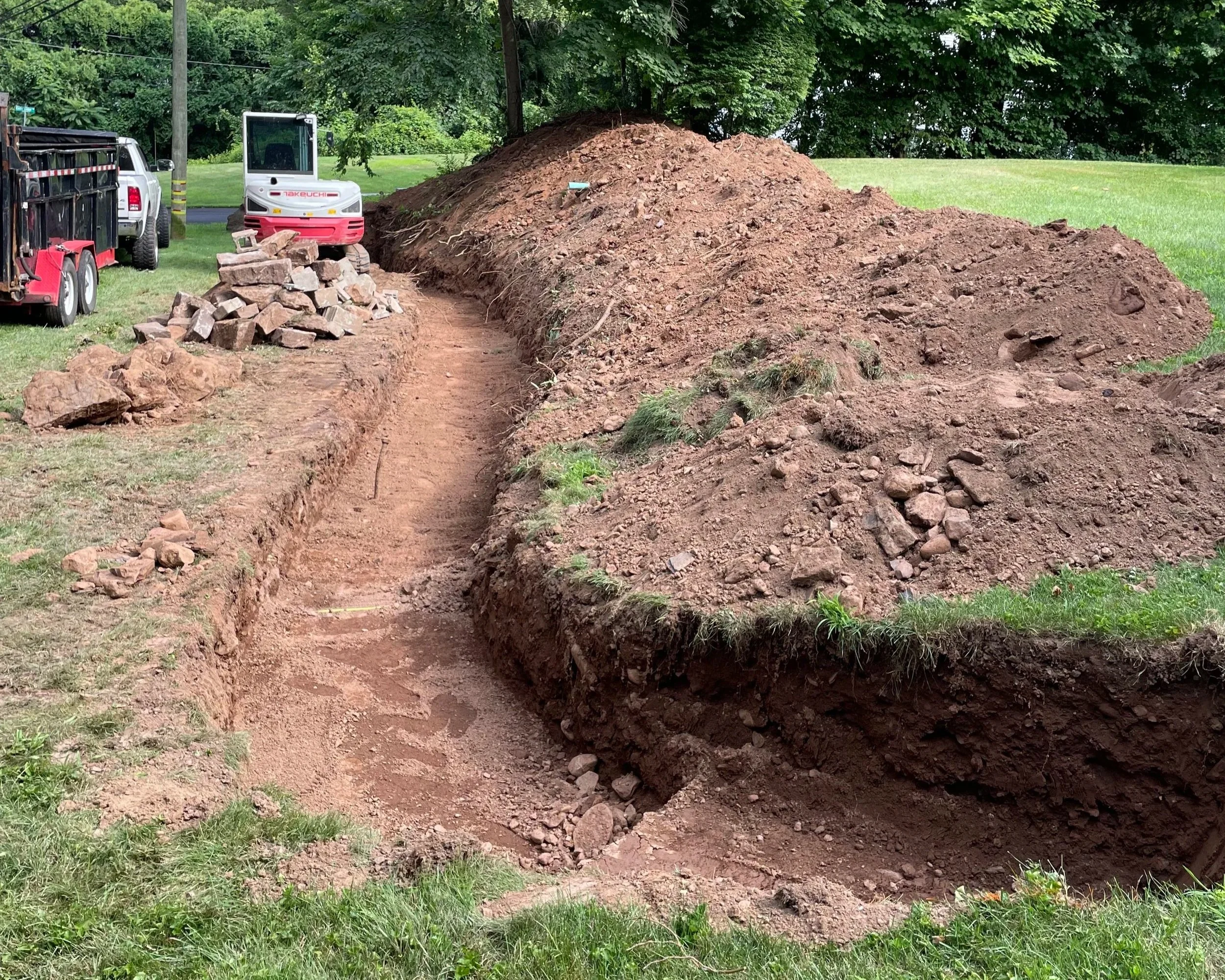A construction site with a dug trench and a large pile of dirt. There are construction vehicles and equipment in the background.