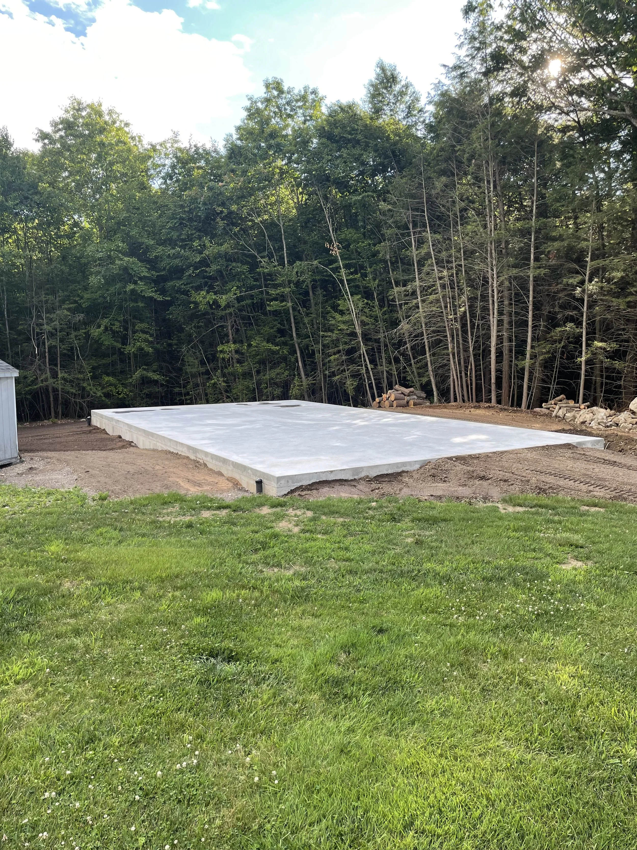 Concrete foundation slab on a grassy area with trees in the background, indicating the early stage of building construction.