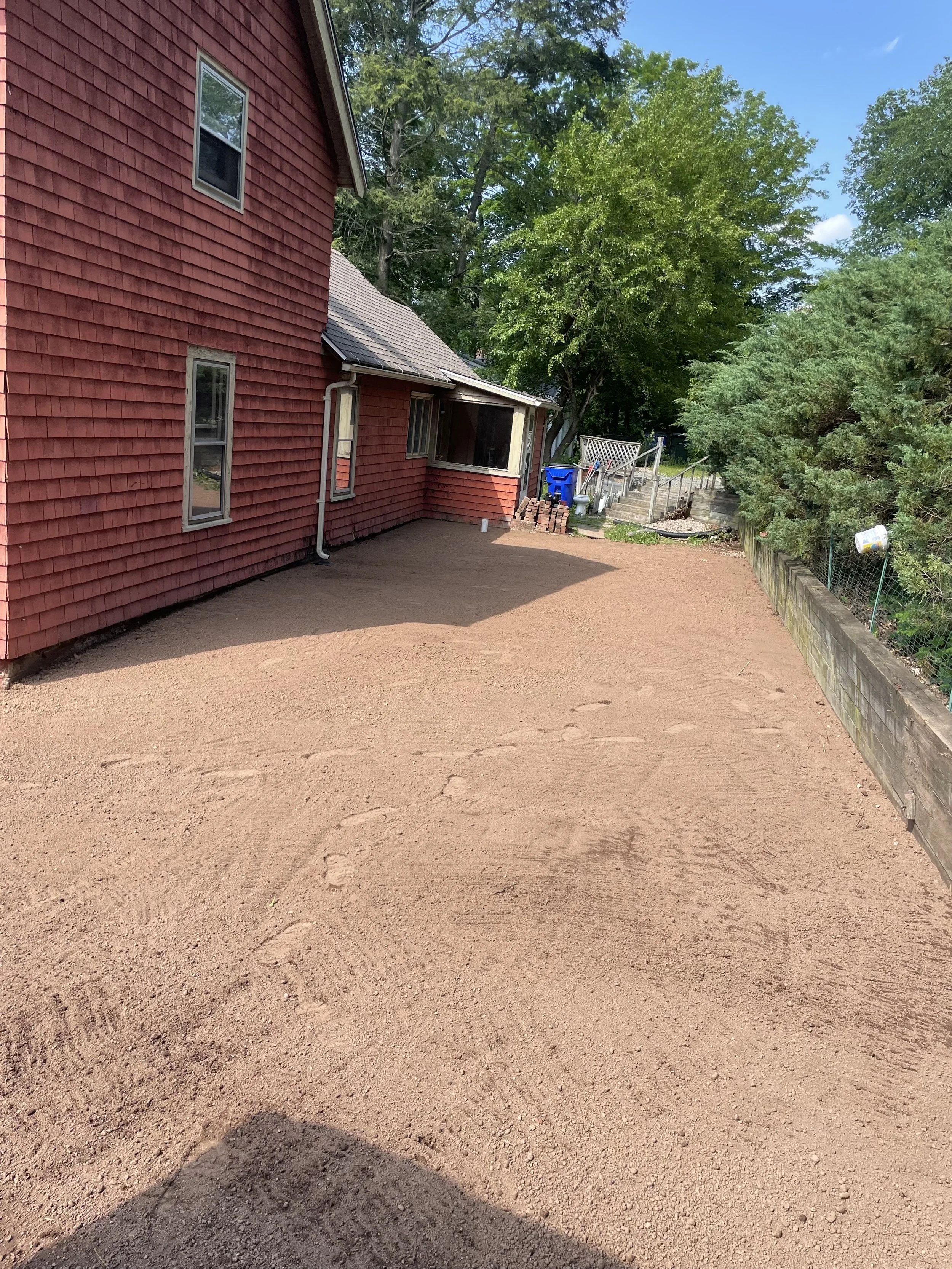 A backyard with a freshly leveled dirt surface next to a red house with multiple windows. The area has some construction materials like bricks, and a fenced garden with a basketball hoop in the background. There are trees and clear blue skies overhead.