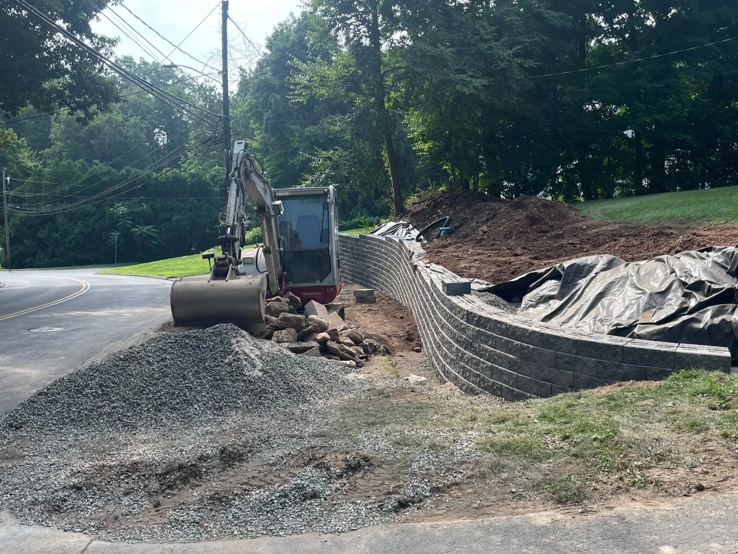Construction site by the side of a curved road with a small excavator pushing rocks into a pile and building a retaining wall. Green trees and grass are visible in the background.