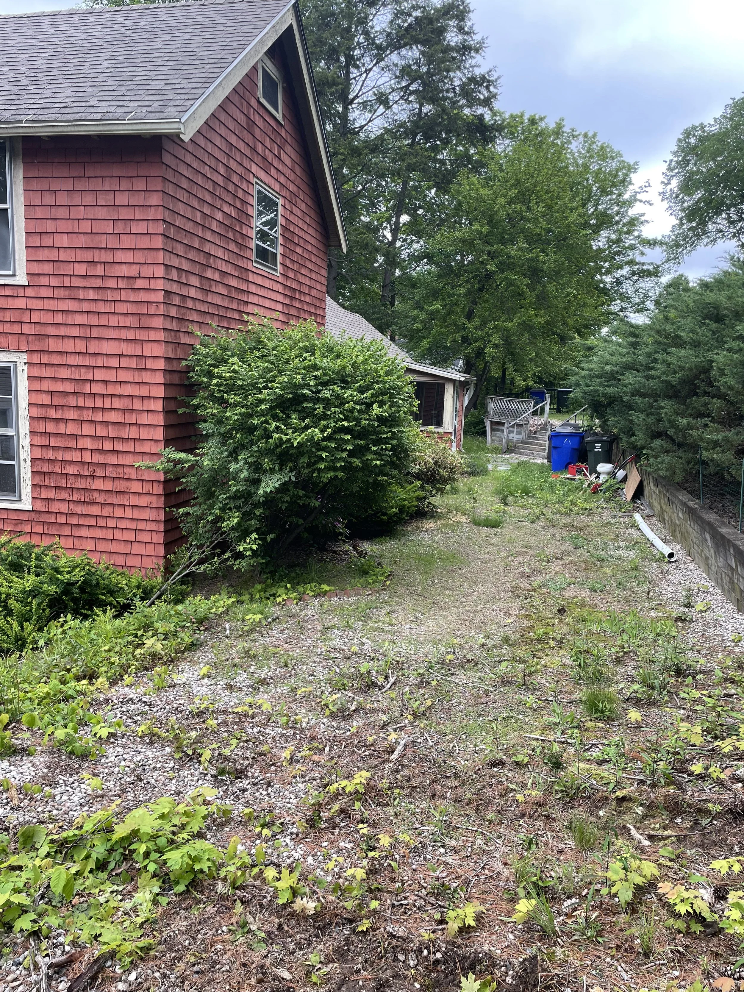 The backyard of a red house with multiple windows and bushes, showing a gravel and dirt ground with some weeds, a few trees, and a small porch with steps leading to the yard. There are trash bins and some fencing along the edge.